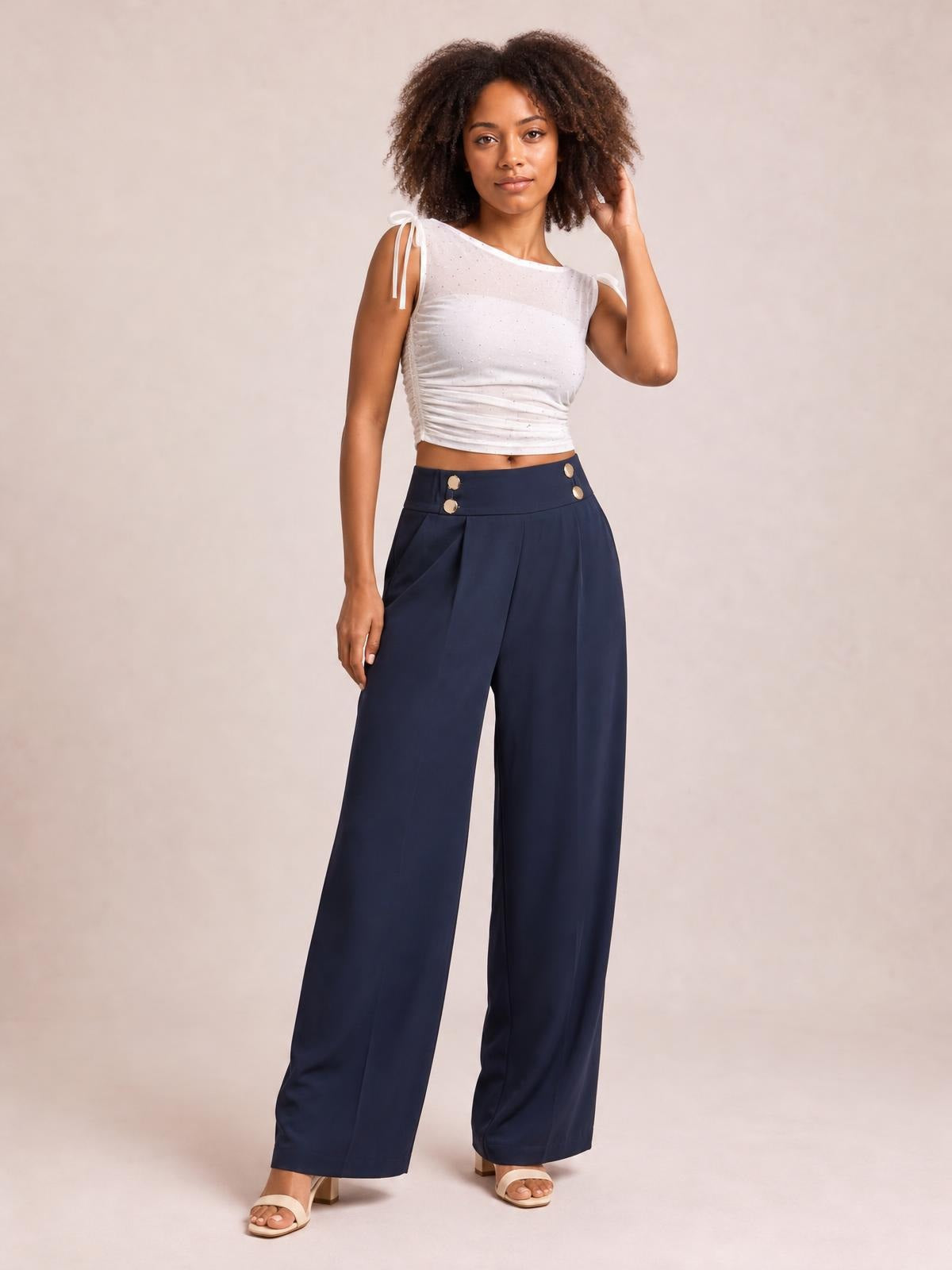 A model poses in navy high-waisted wide-leg pants with pleated fronts, a structured waistband, and visible decorative buttons.