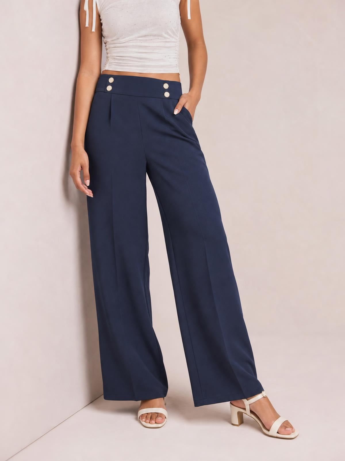Model wearing navy high-waisted wide-leg pants with front pleats, button details, and side pockets, standing with one hand in a pocket.