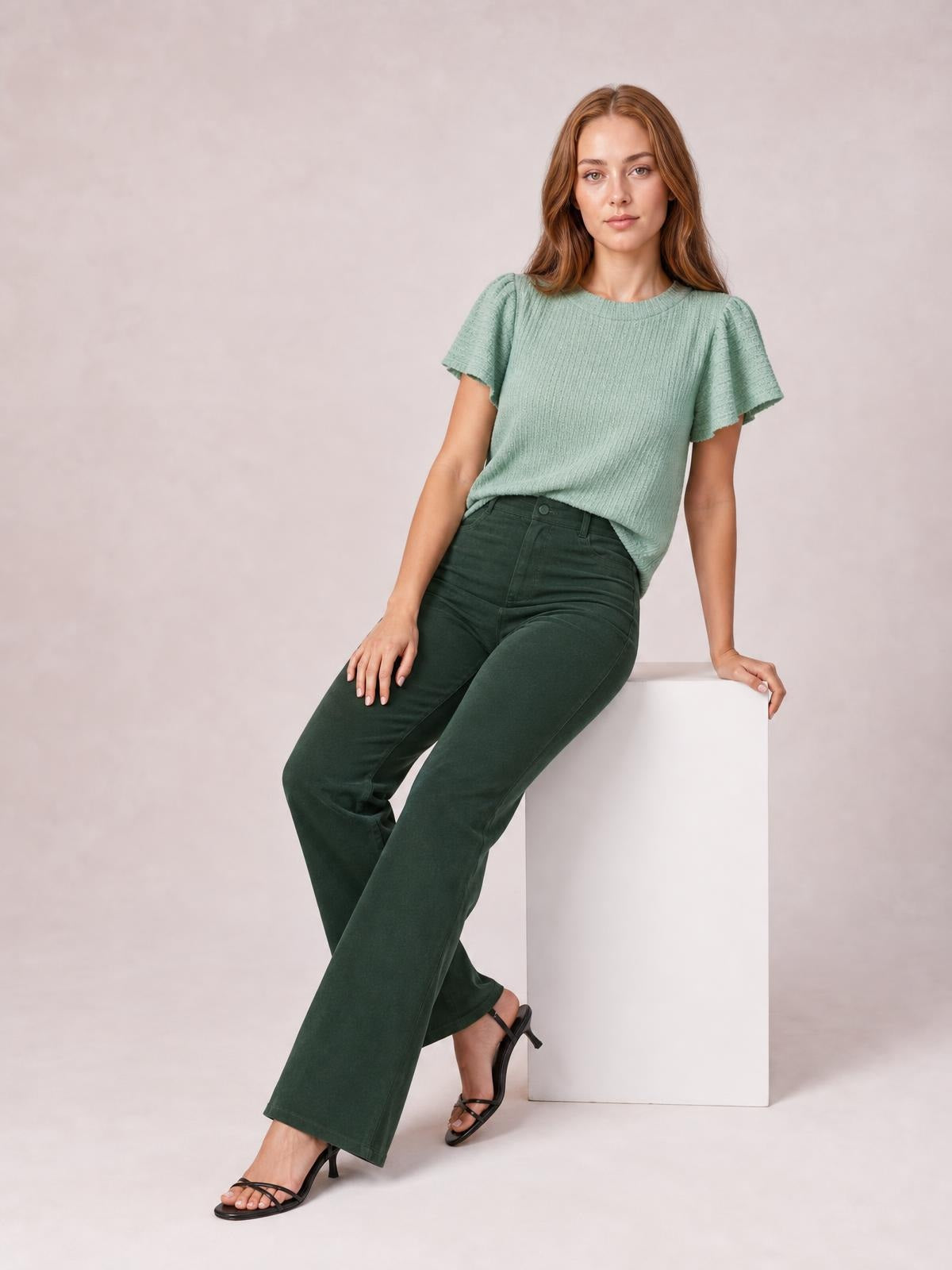 Model seated on a white block wears dark green high-waist bootcut pants with a fitted silhouette, front button closure, belt loops, and flared hems.