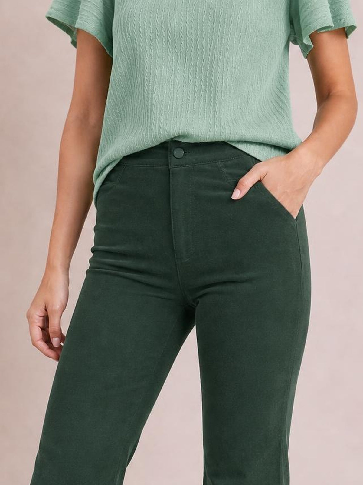A model wears dark green high-waisted pants with a front button closure, fitted hips and thighs, and one hand resting in the angled side pocket.