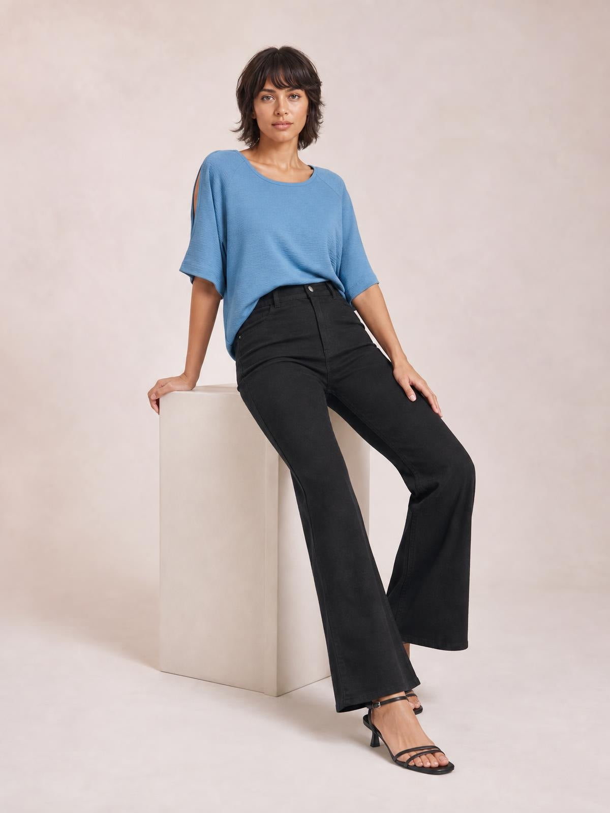 A seated model wears black high-waisted bootcut pants with a fitted waist, belt loops, front button closure, and flared legs.