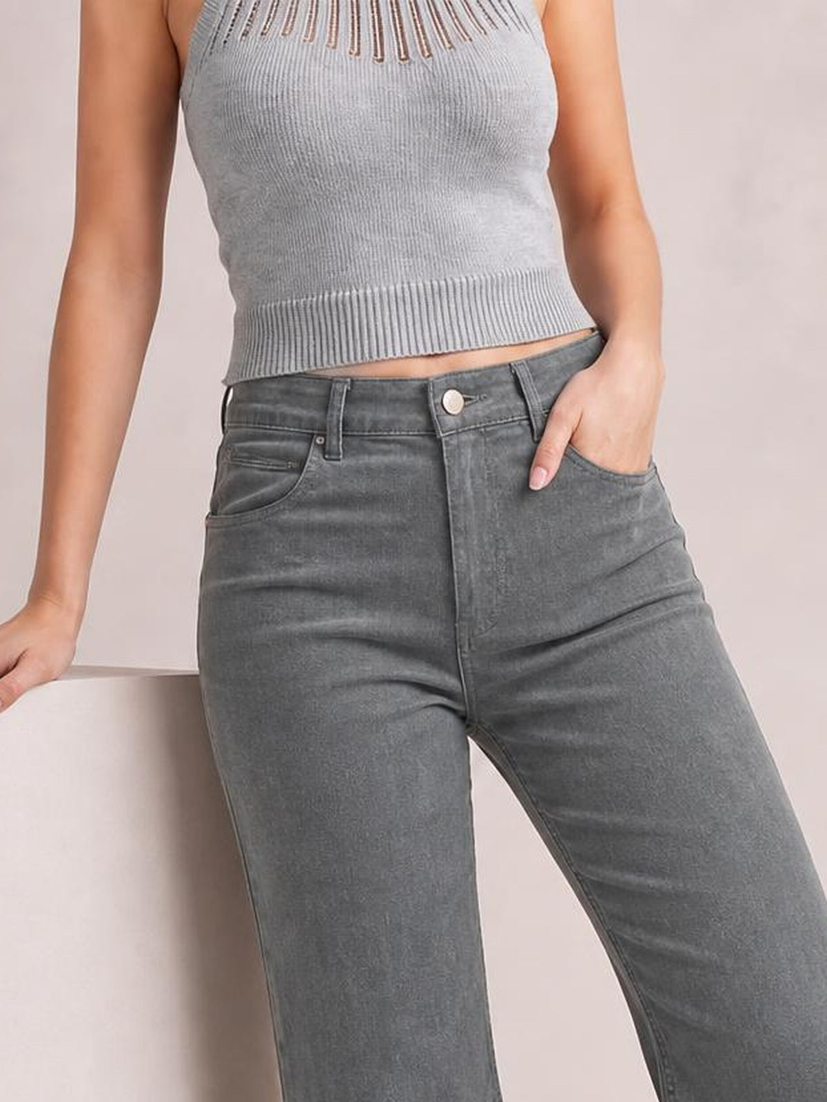 A model wears gray high-waist bootcut pants with a structured waistband, belt loops, button closure, front pockets, and a relaxed pose with one hand in a pocket.