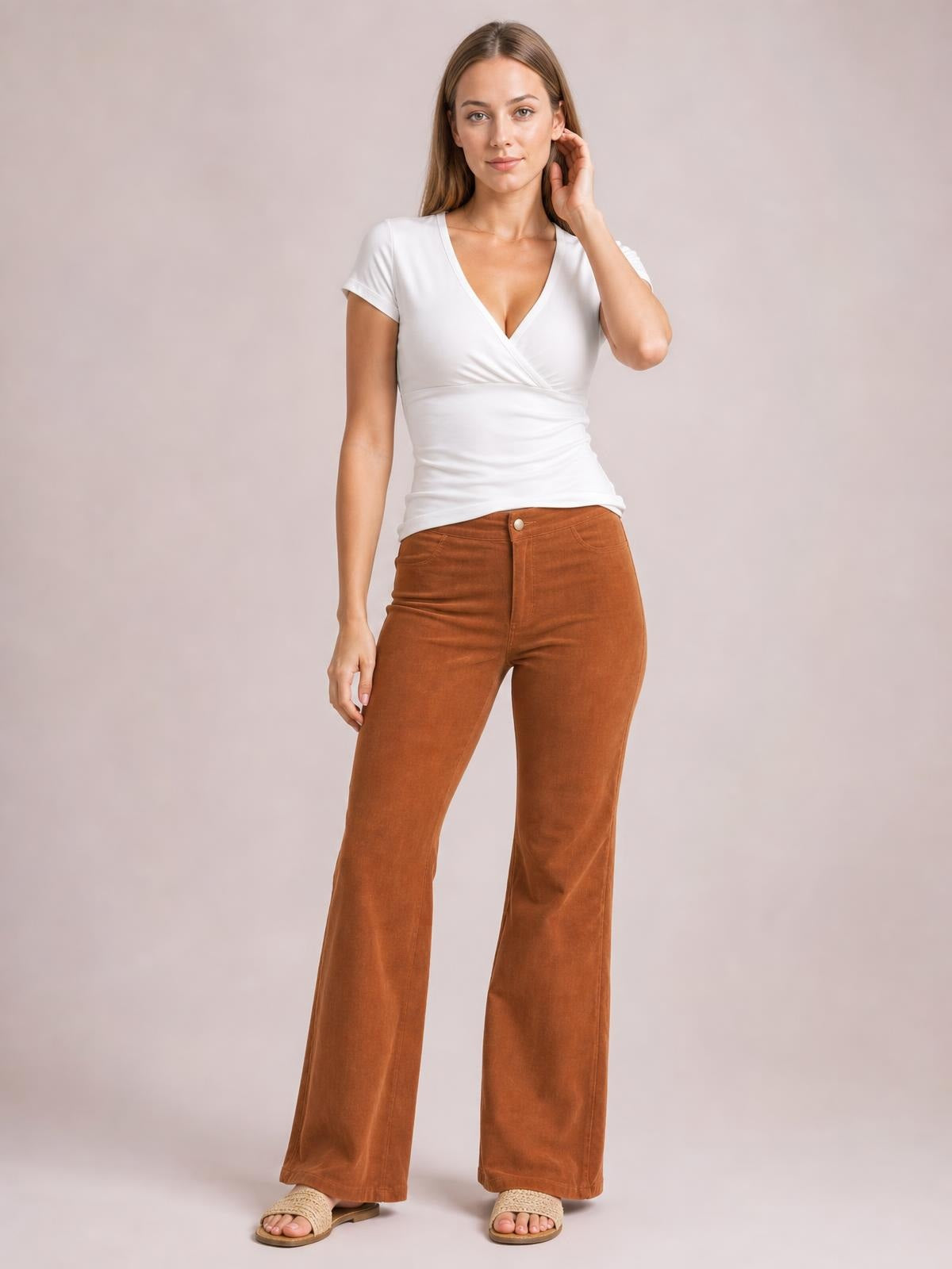 A model stands in brown high-waist bootcut pants with a button closure, front pockets, fitted hips, and gently flared legs, one hand raised to her hair.