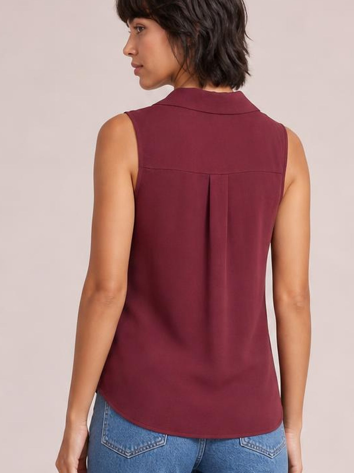 A model turns to show the burgundy Johnny collar button-up tank’s sleeveless cut, relaxed silhouette, back yoke seam, center pleat, and curved hem.
