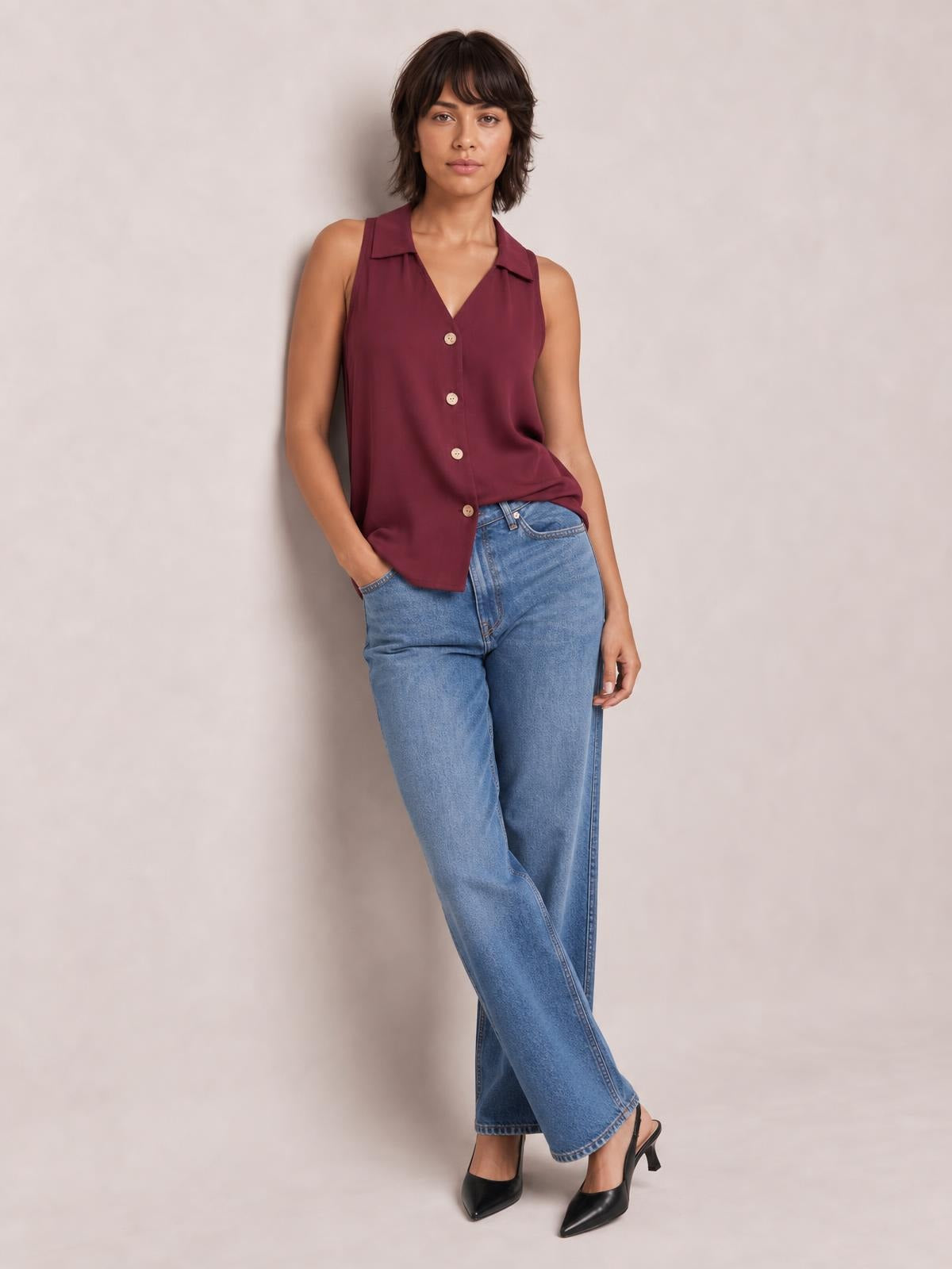 Model poses in a burgundy Johnny collar button-up tank with a sleeveless, relaxed silhouette and front buttons, paired with jeans.