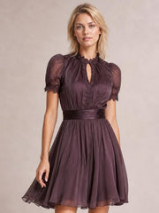 A model stands with one hand on the skirt, wearing a plum A-line dress with puff sleeves, lace-trimmed neckline and cuffs, keyhole front, and gathered waist.