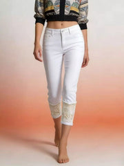 A model wears white lace-trimmed capri jeans with a fitted silhouette, button closure, front pockets, and detailed lace cuffs while standing with one leg crossed.