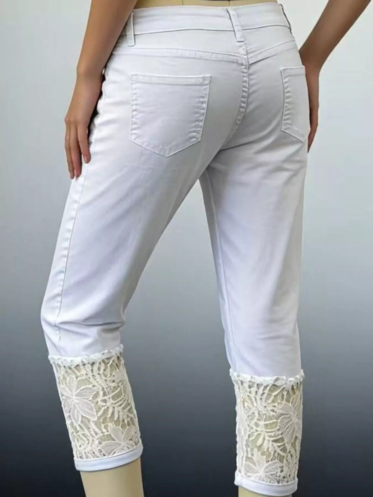 Model standing with hands at her sides wears white lace-trimmed capri jeans with a fitted silhouette, button waistband, belt loops, back pockets, and lace cuffs.