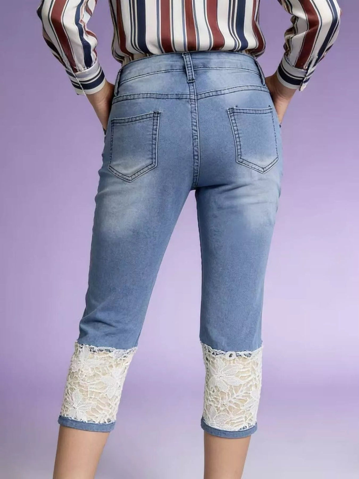 Model shown from behind wears light blue lace-trimmed capri jeans with a fitted straight-leg silhouette, rear patch pockets, and white lace panels at the cropped cuffs.
