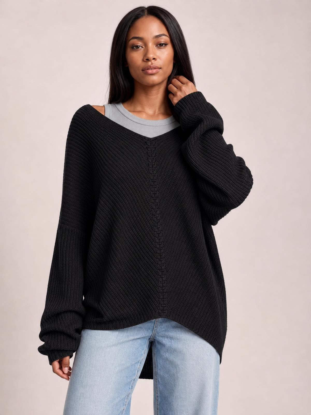 A model wears a black lace-up V-neck long-sleeve sweater with ribbed knit texture and an oversized relaxed silhouette, touching her hair while standing in jeans.