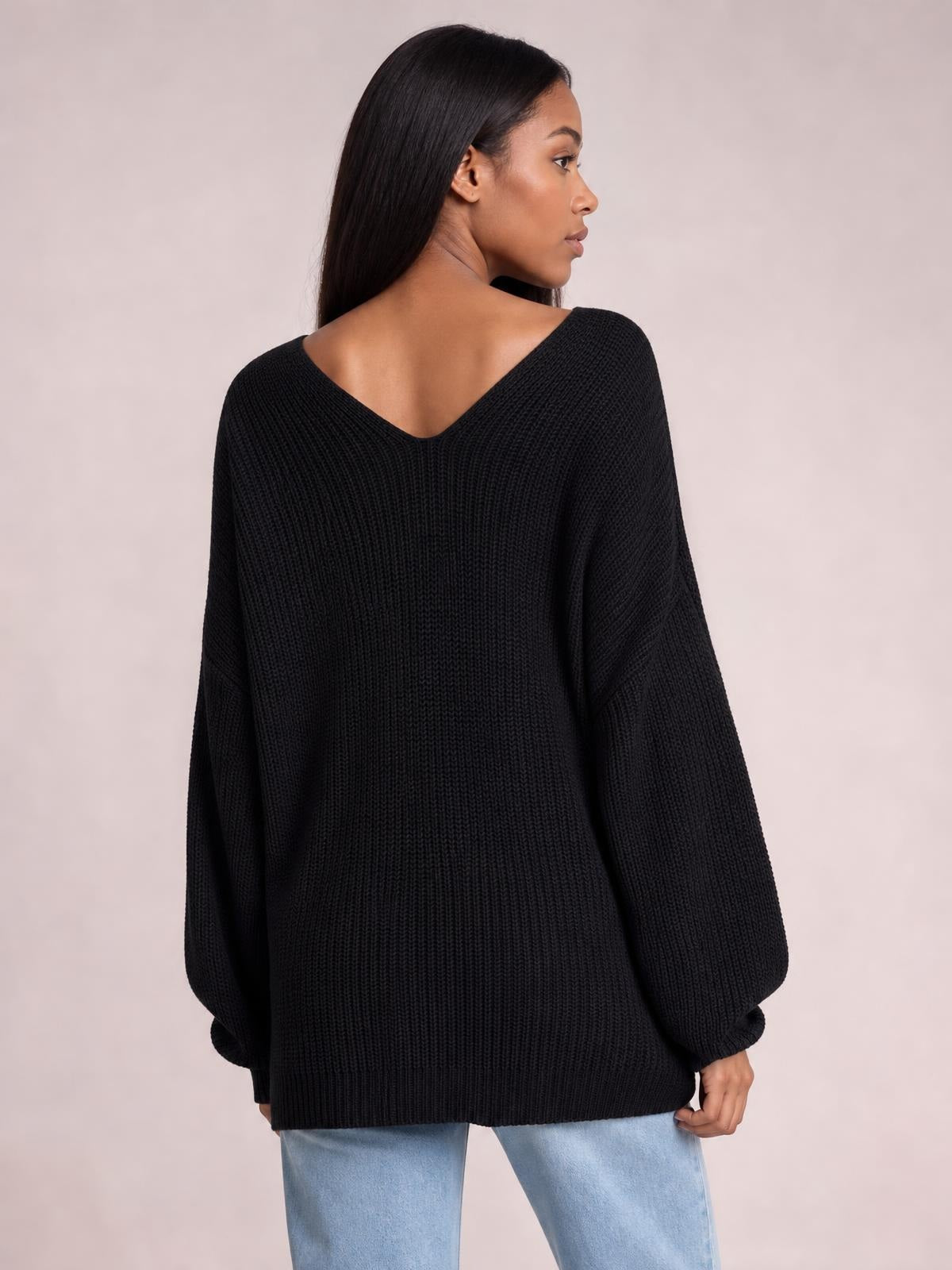 A model looks over her shoulder wearing a black long sleeve sweater with a deep V-neck back, ribbed knit construction, and a relaxed oversized silhouette.