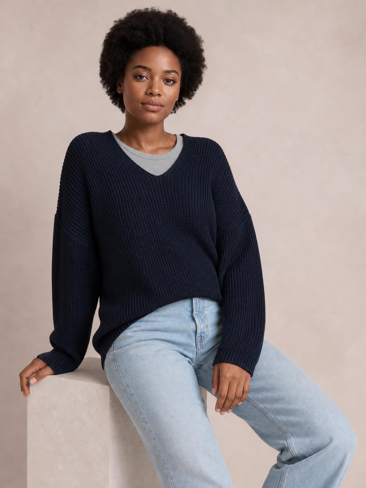 A model sits in a relaxed dark navy ribbed V-neck sweater with long sleeves and a loose silhouette, layered over a gray top and light-wash jeans.