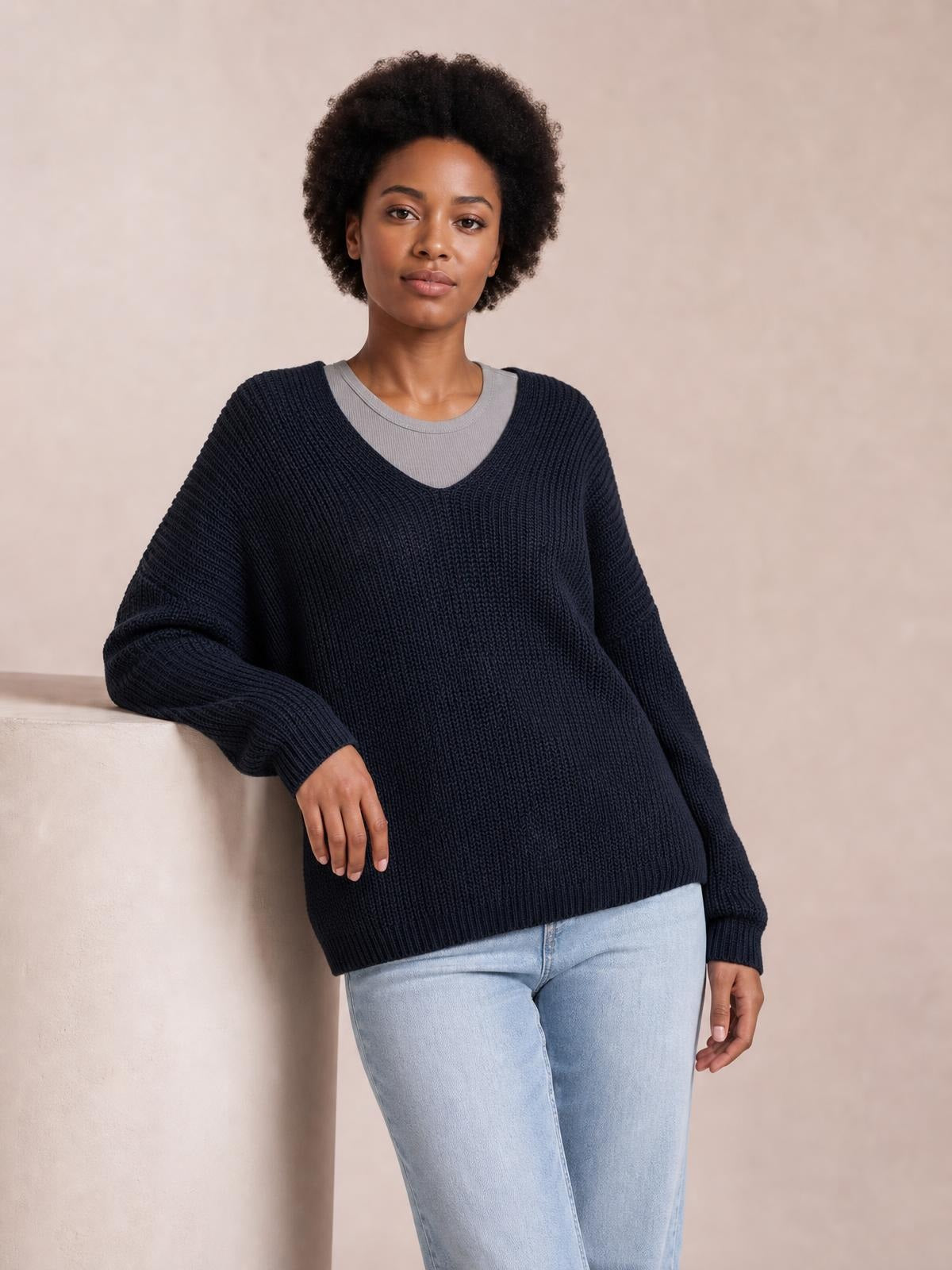Model wears a dark navy V-neck long-sleeve sweater with ribbed knit construction and a relaxed, slightly oversized silhouette while leaning casually.