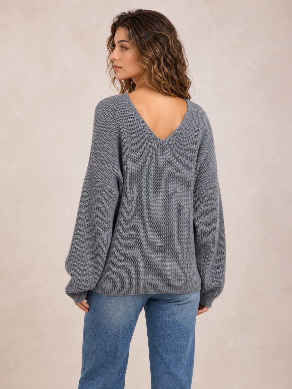 Model turned slightly to the side, showcasing a gray knit sweater with a deep V-neck back, long sleeves, relaxed oversized silhouette, and ribbed cuffs.