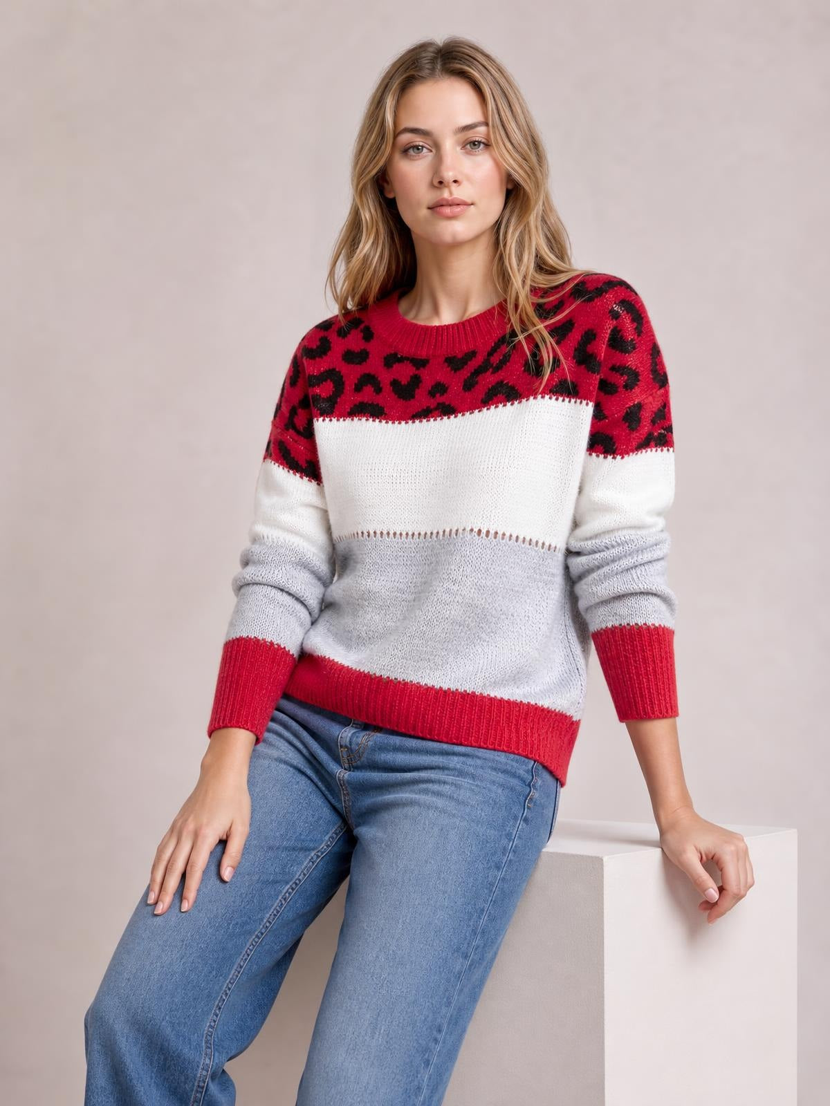 Model seated in a leopard color block long sleeve sweater with red, white, and gray sections, ribbed crew neck, cuffs, and hem, styled with jeans.