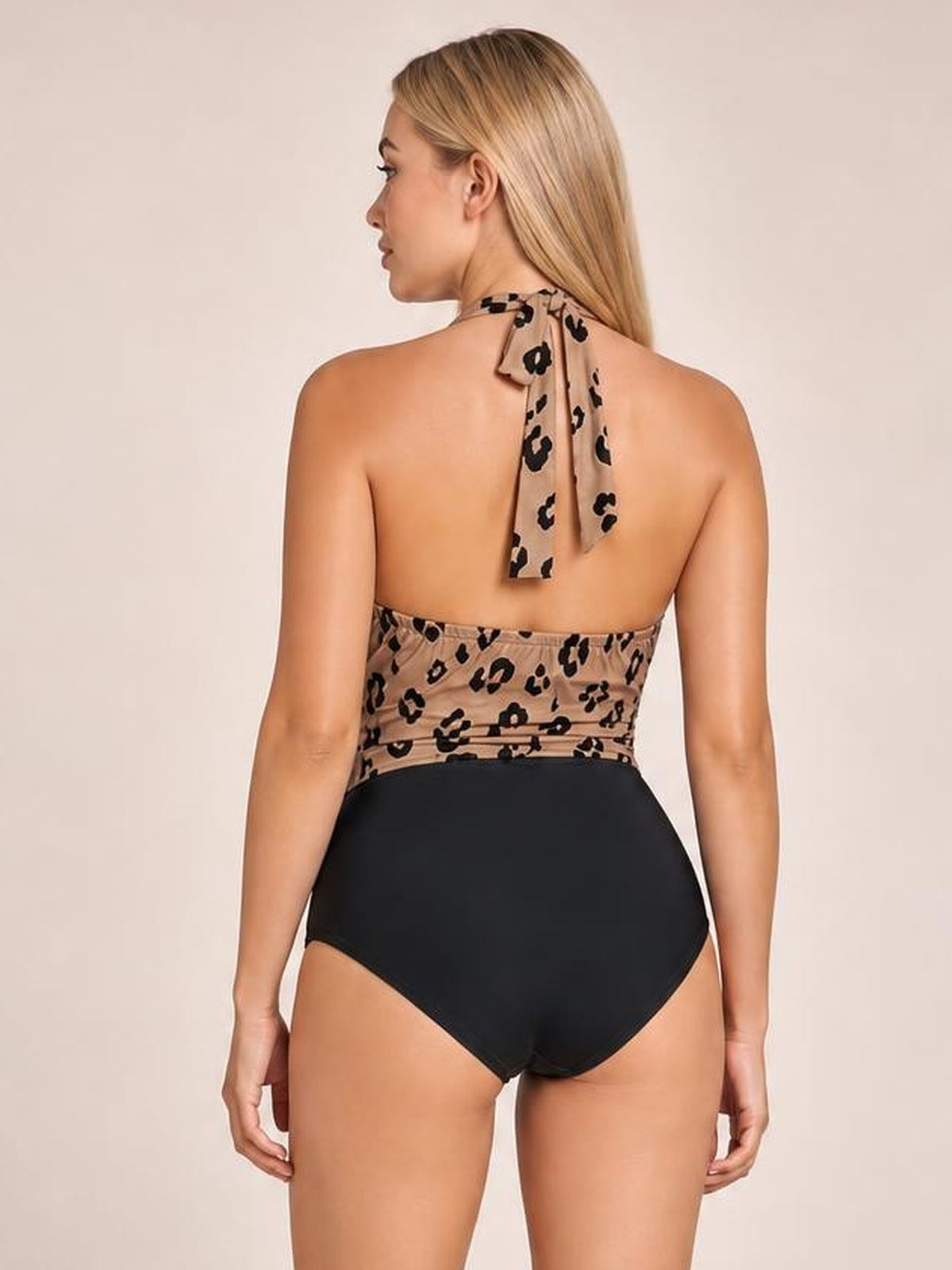 Rear view of a model standing in a leopard halter neck one-piece swimsuit with a tied neck, open back, fitted leopard-print bodice, and black high-waisted bottom.