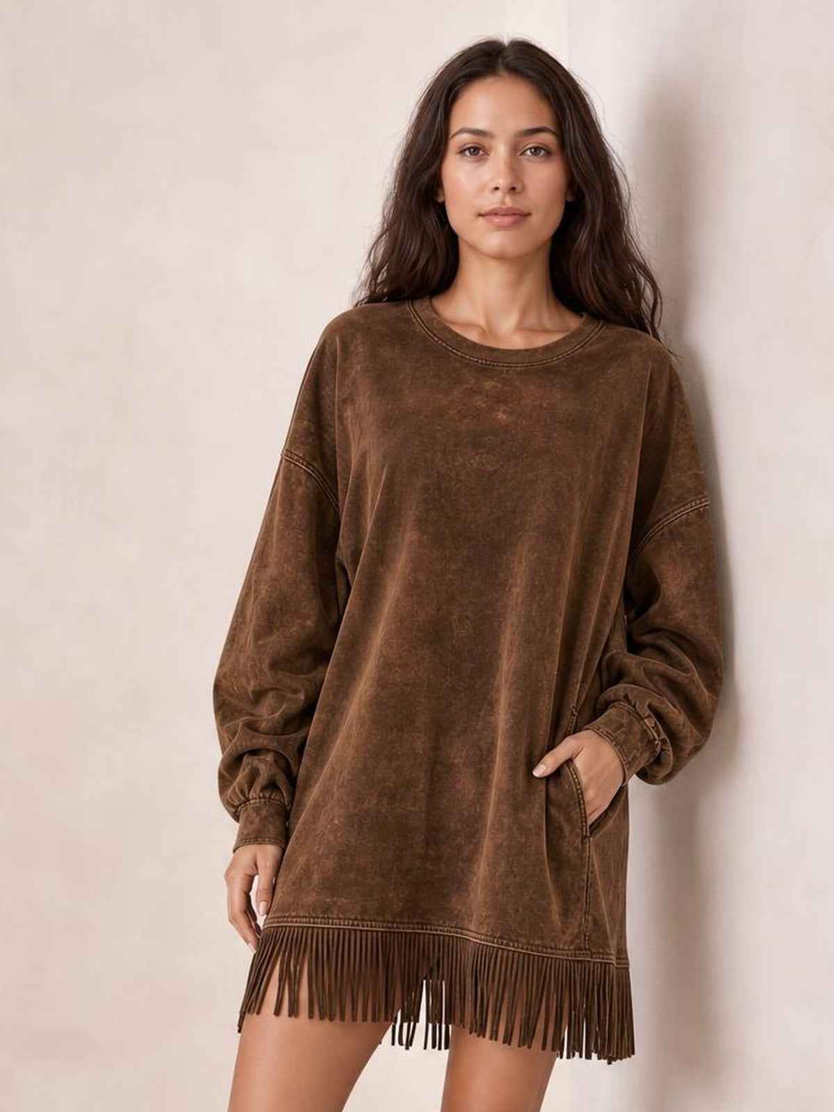 Model wears a relaxed brown long-sleeve mini dress with a mock neck, oversized silhouette, side pockets, seam detailing, and a fringed hem.