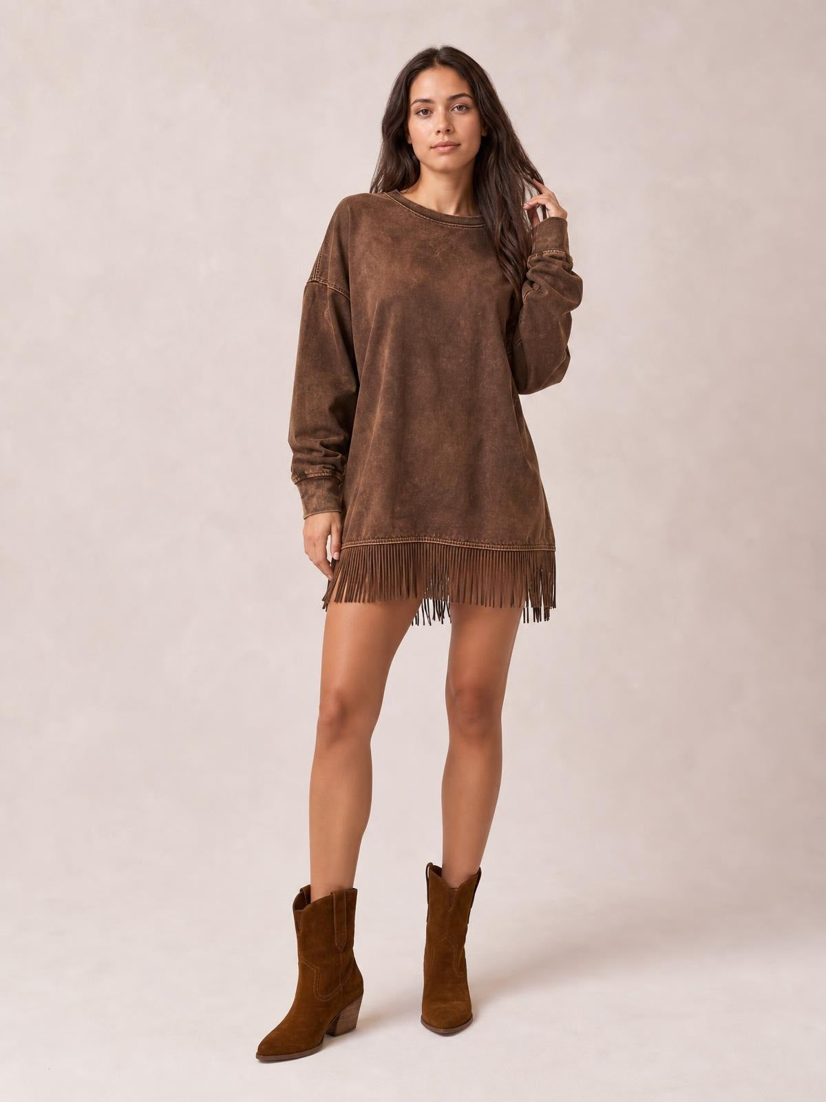 Model poses in a brown long-sleeve mini dress with a relaxed straight silhouette, round neckline, seam details, and long fringe along the hem, touching her hair.