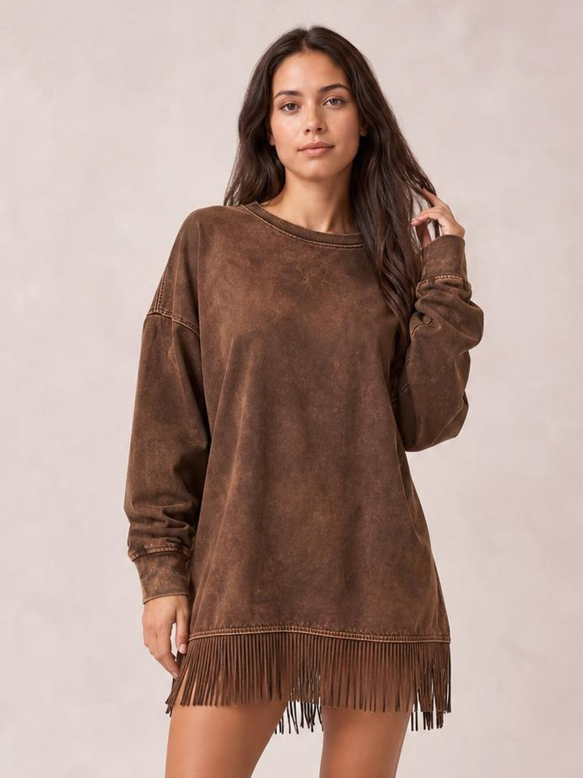 A model poses in a relaxed brown long-sleeve mini dress with a high neckline, loose silhouette, visible seam detailing, and a fringed hem.