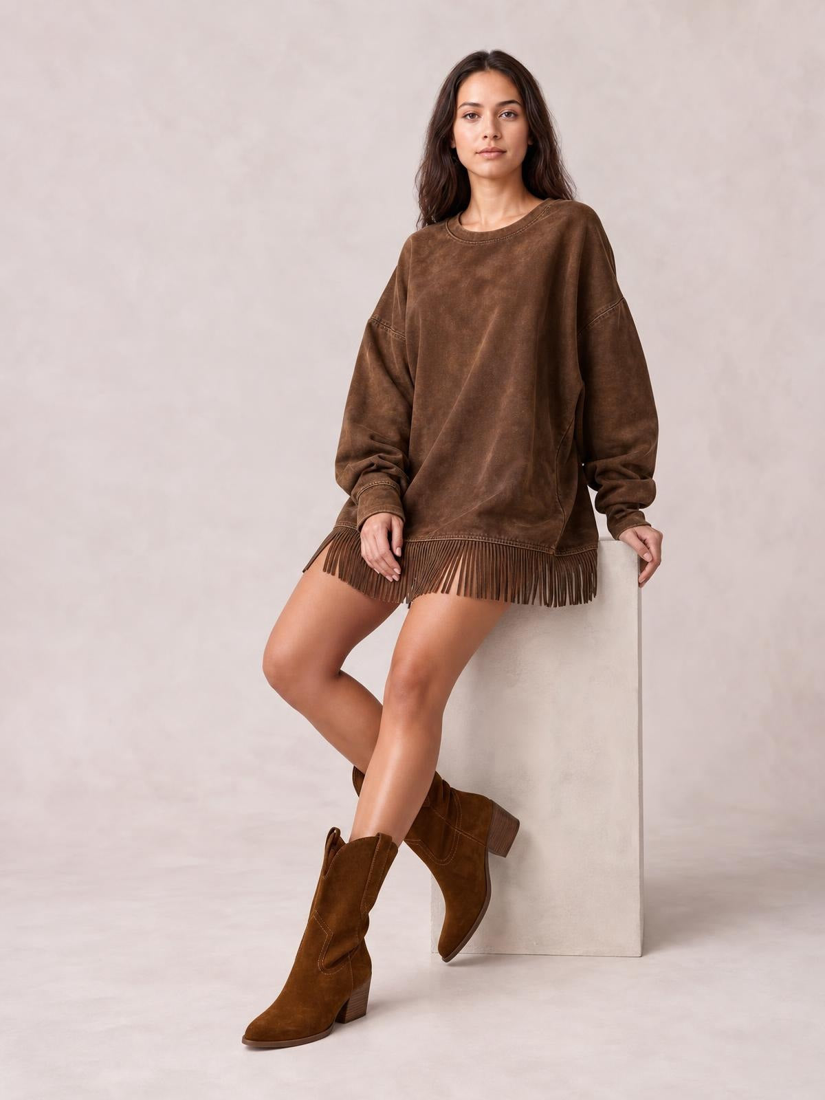 A model sits with crossed legs in a relaxed brown long-sleeve mini dress featuring dropped shoulders, exposed seam details, and a fringed hem.