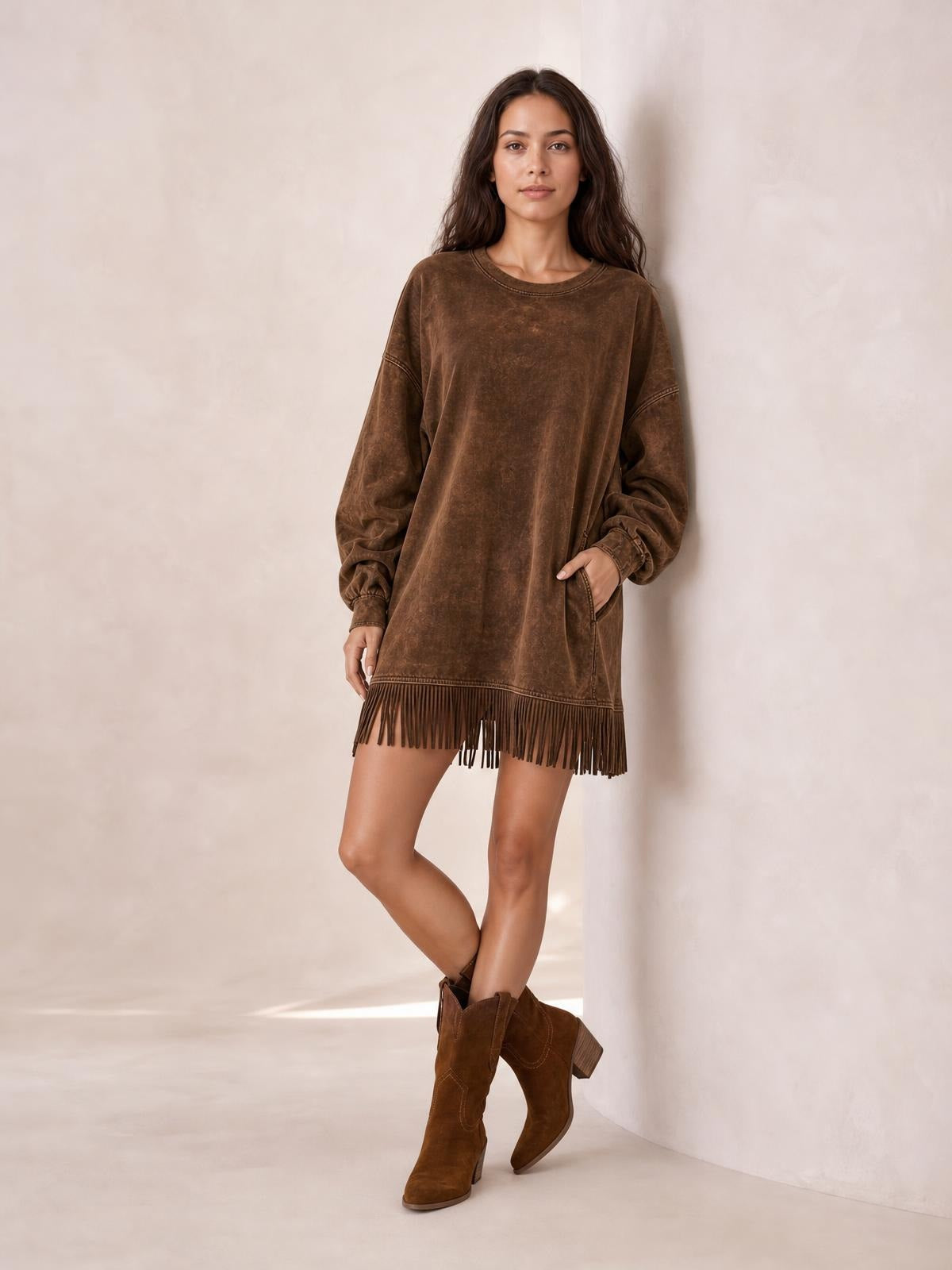 A model poses with crossed legs in a relaxed brown long-sleeve mini dress featuring a loose silhouette, side pockets, and a fringe-trimmed hem.