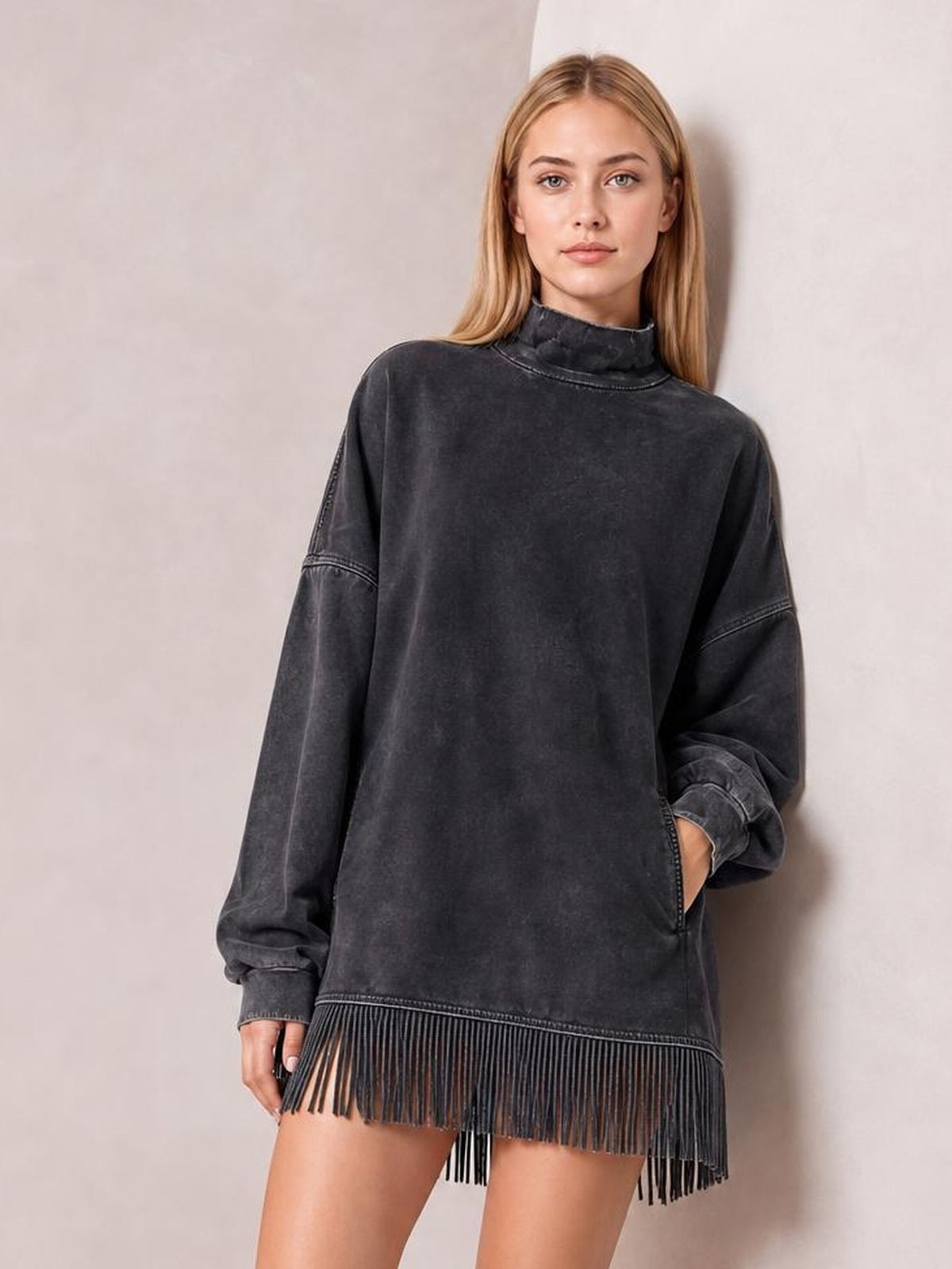 Model poses in a charcoal mock neck long sleeve mini dress with an oversized silhouette, side pocket, cuffed sleeves, and fringed hem.