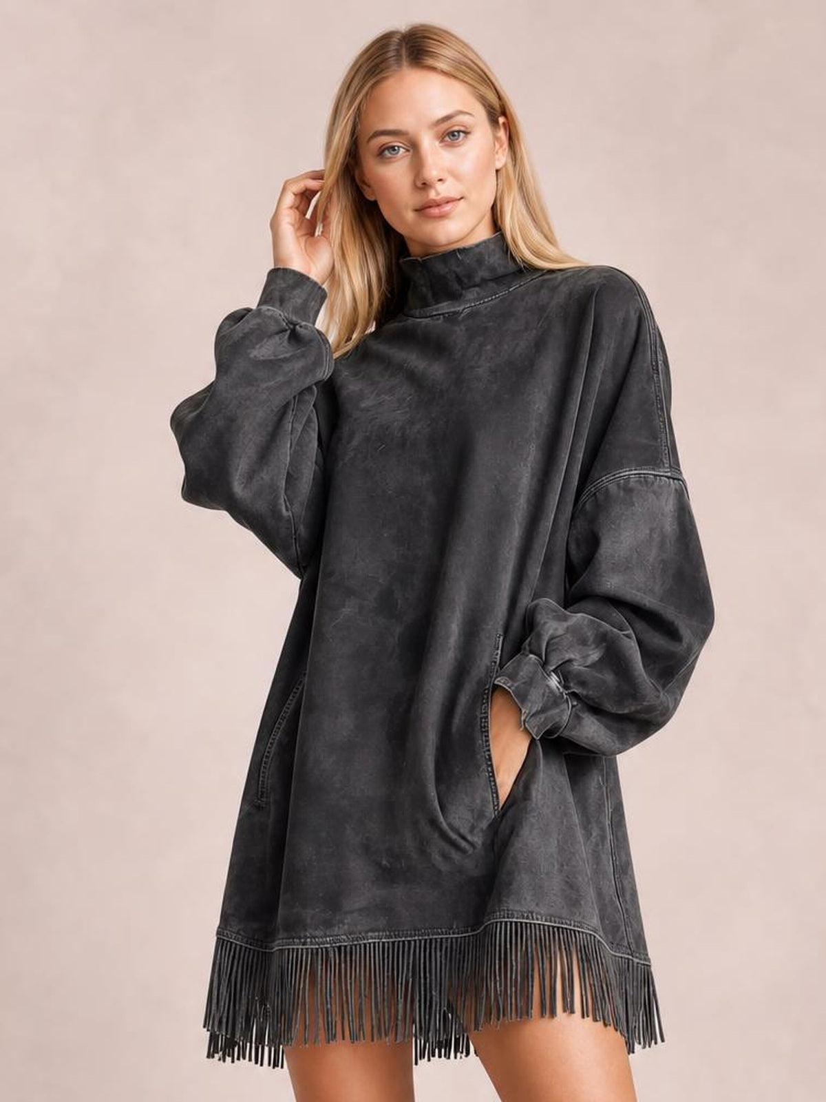 Model poses in a charcoal mock neck mini dress with long balloon sleeves, seam detailing, side pockets, a relaxed silhouette, and fringed hem.
