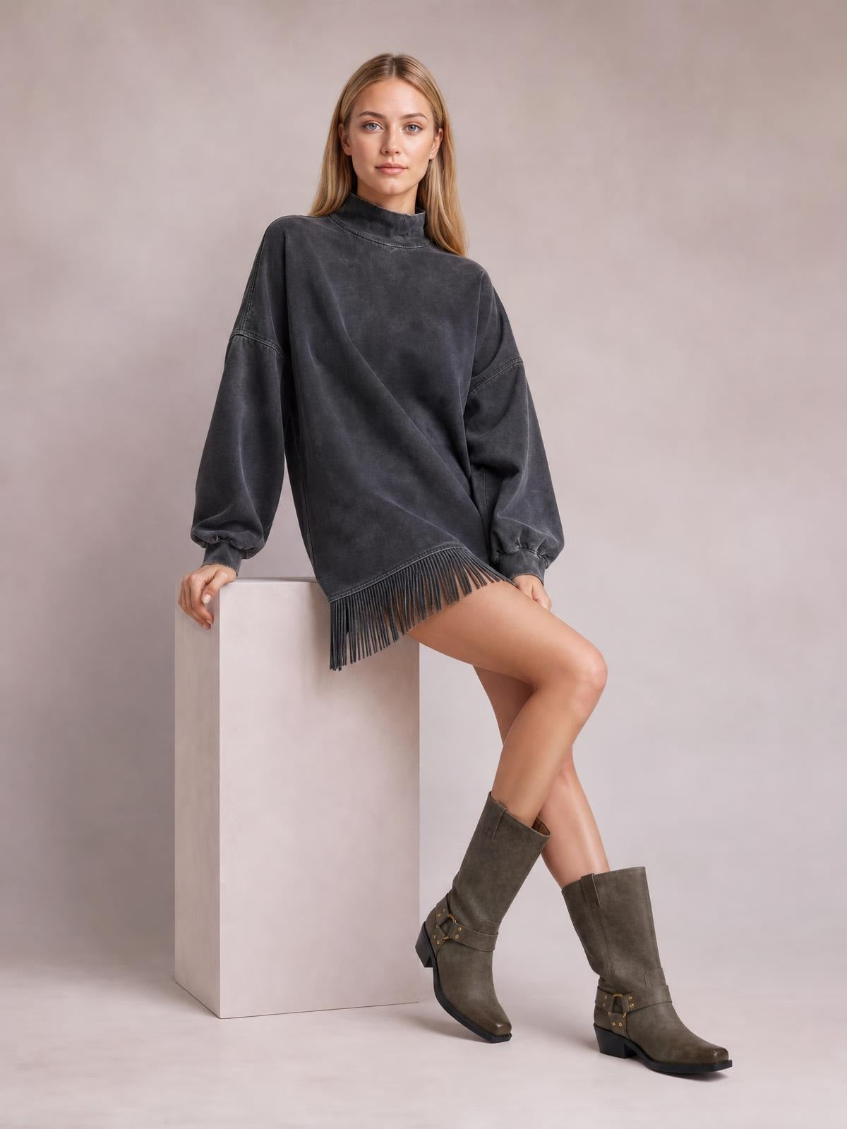 A model leans against a cube wearing a charcoal gray mock neck mini dress with long sleeves, an oversized silhouette, seamed construction, and fringed hem.