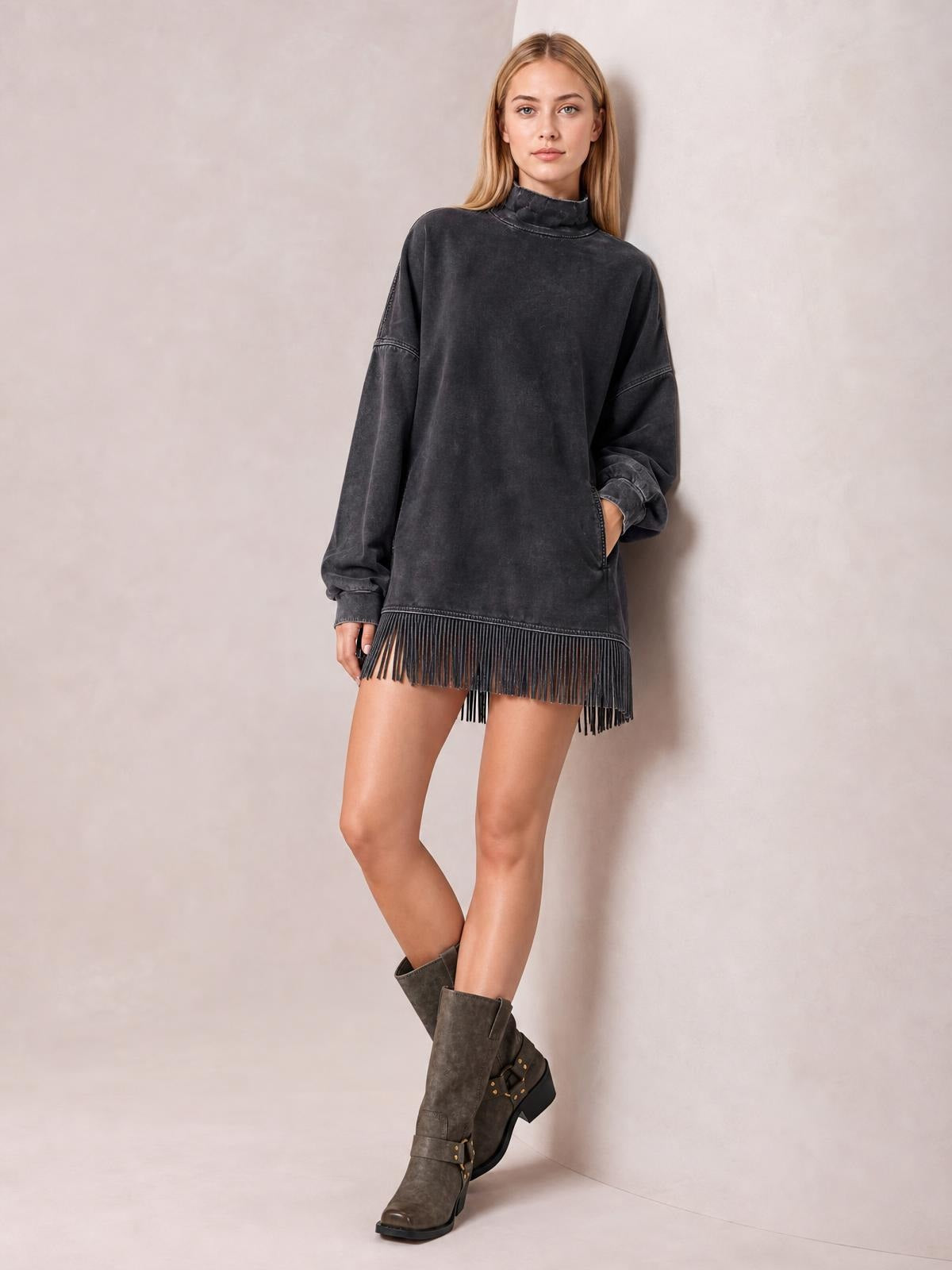 A model poses in a charcoal mock-neck mini dress with long sleeves, an oversized straight silhouette, side pockets, and a fringed hem.