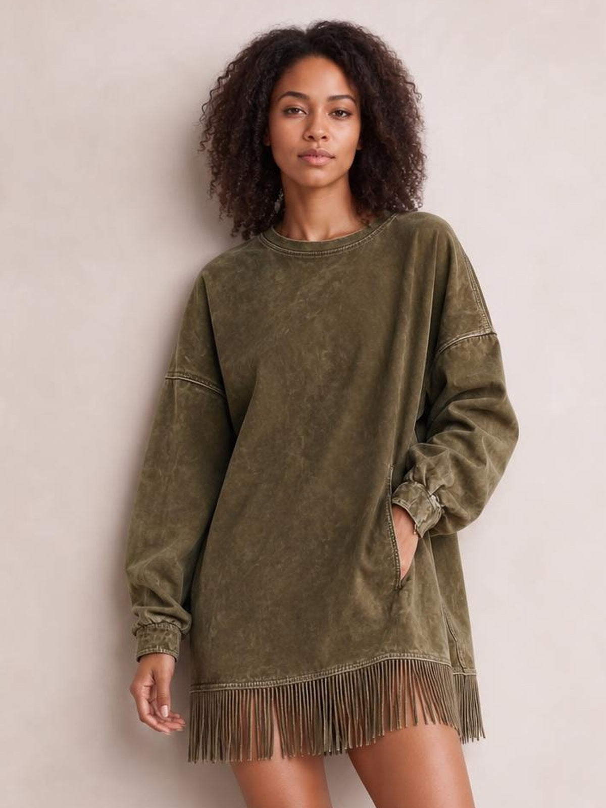 A model poses in an olive green mock neck long sleeve mini dress with a relaxed silhouette, side pocket, cuffed sleeves, and dramatic fringed hem.