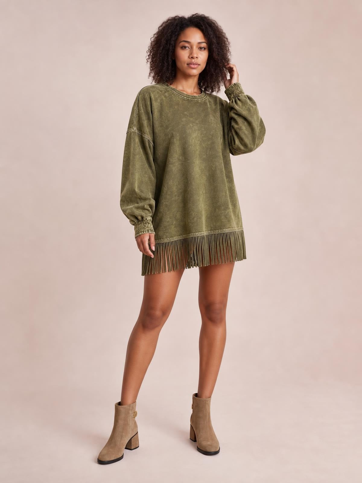 A model poses in an olive-green long-sleeve mini dress with a mock neckline, relaxed silhouette, seam details, and a fringe-trimmed hem.