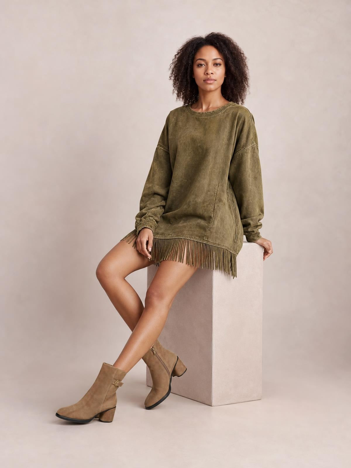 Model seated in an olive mock-neck long-sleeve mini dress with a relaxed silhouette, side pockets, and a fringed hem.