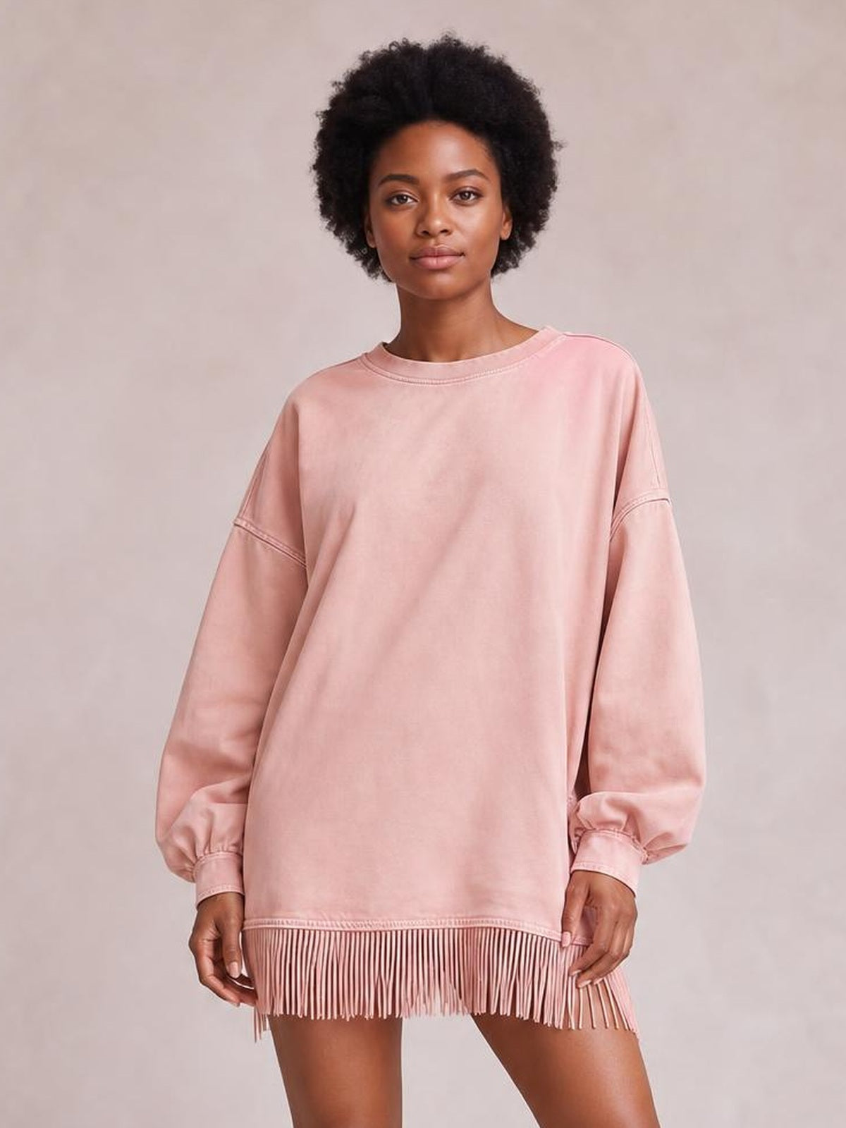 A model stands holding the hem of a pink long-sleeve mini dress with a relaxed straight silhouette, round neckline, cuffed sleeves, and long fringe trim.