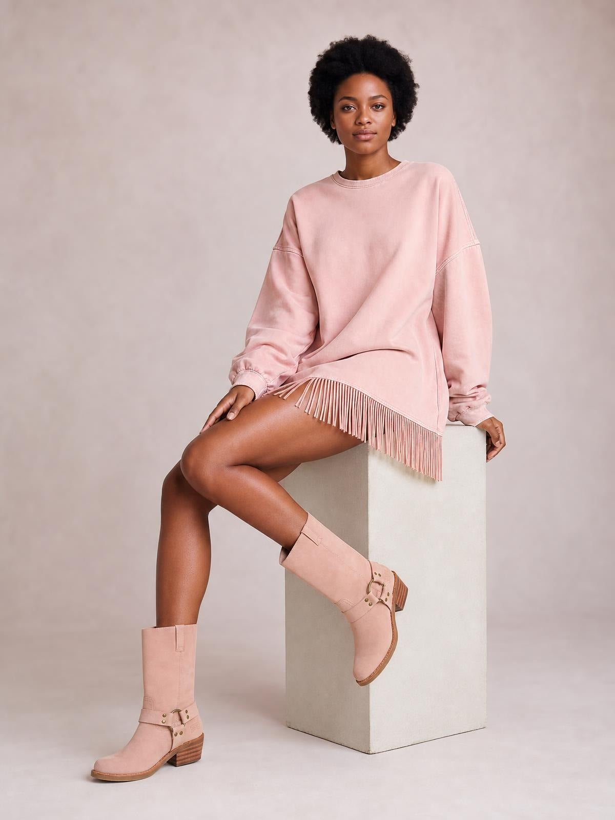 A seated model wears a pink long-sleeve mini dress with a mock neck, relaxed silhouette, seam details, and a dramatic fringed hem.