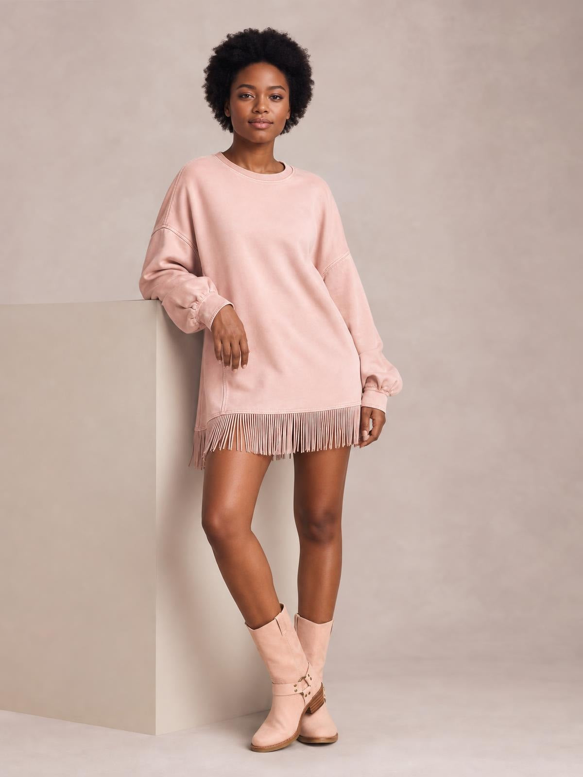 A model leans against a ledge wearing a relaxed pink long-sleeve mini dress with seam details, gathered cuffs, and a long fringed hem.