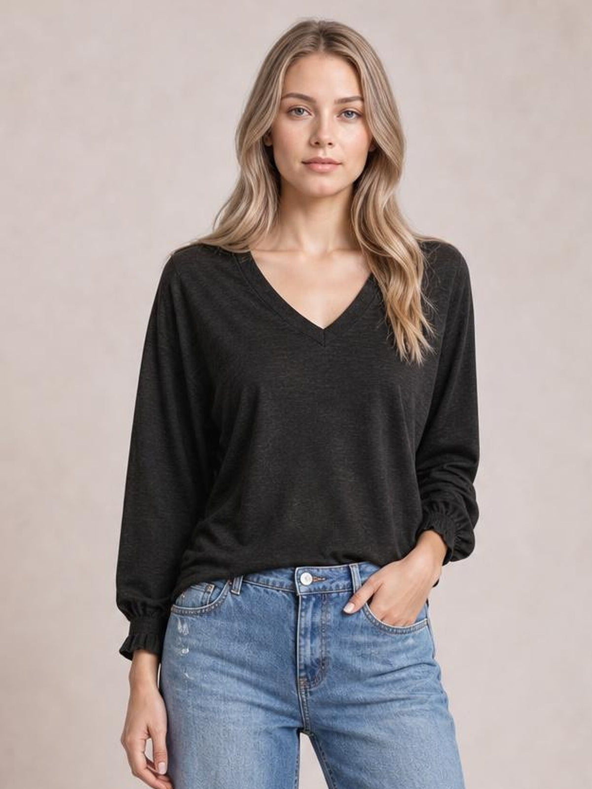 Model wears a black notched long sleeve T-shirt with a relaxed silhouette, V-neckline, and subtly gathered ruffle cuffs, posing with one hand in her jeans pocket.
