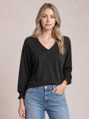 Model wears a black notched long sleeve T-shirt with a relaxed silhouette, V-neckline, and subtly gathered ruffle cuffs, posing with one hand in her jeans pocket.