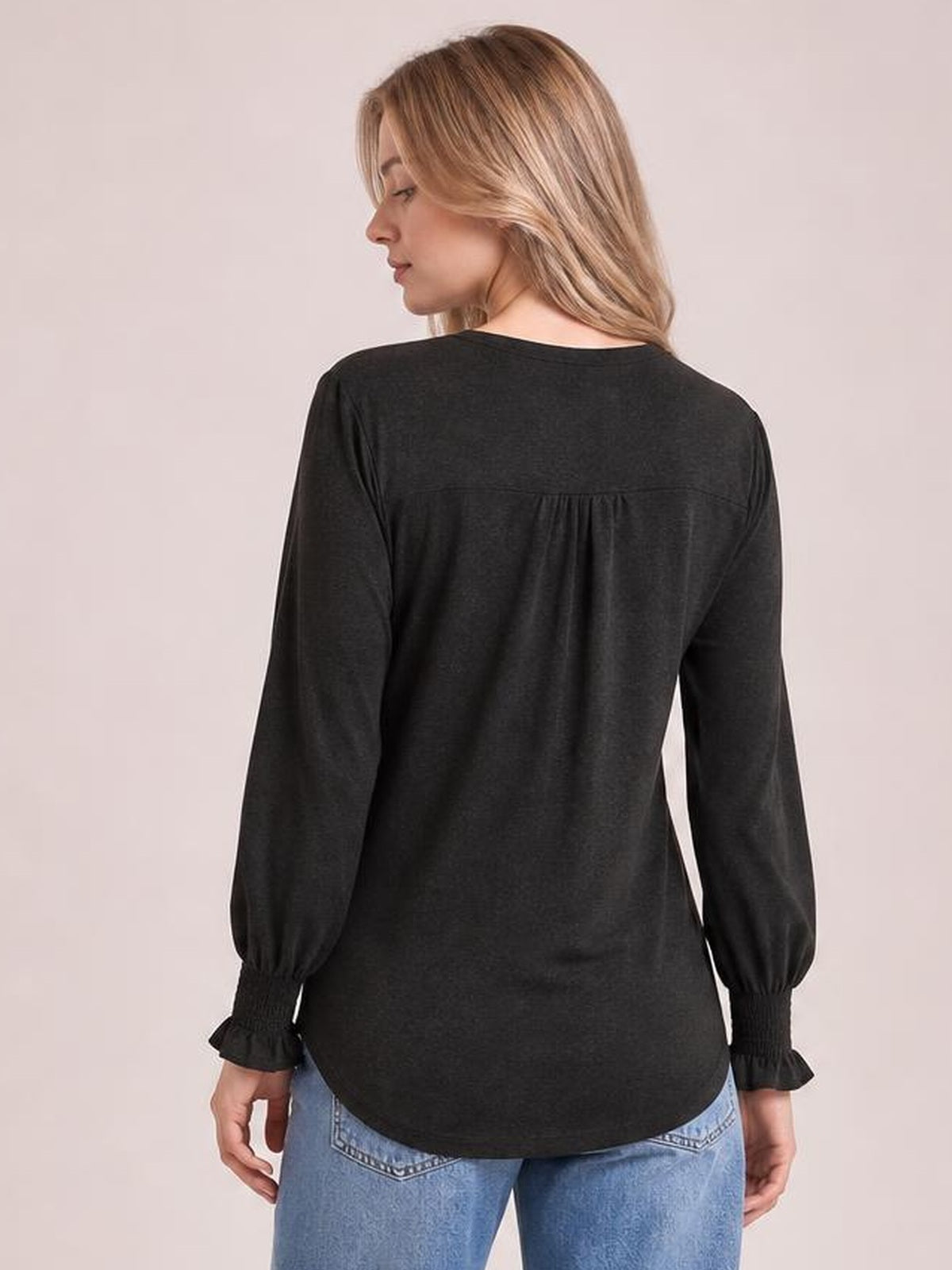 A model stands in a back view wearing a black long sleeve t-shirt with a relaxed silhouette, gathered back yoke, and ruffled cuffs.