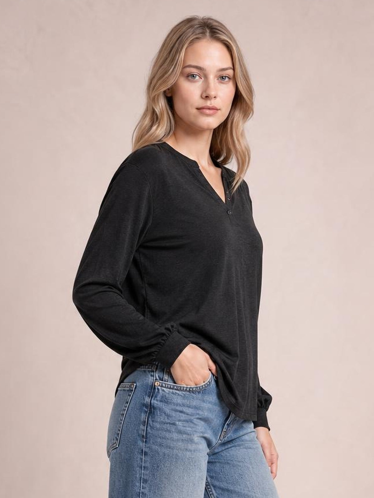 Model wears a black notched long sleeve T-shirt with a relaxed silhouette, buttoned neckline, and ribbed cuffs while posing with one hand in her jeans pocket.