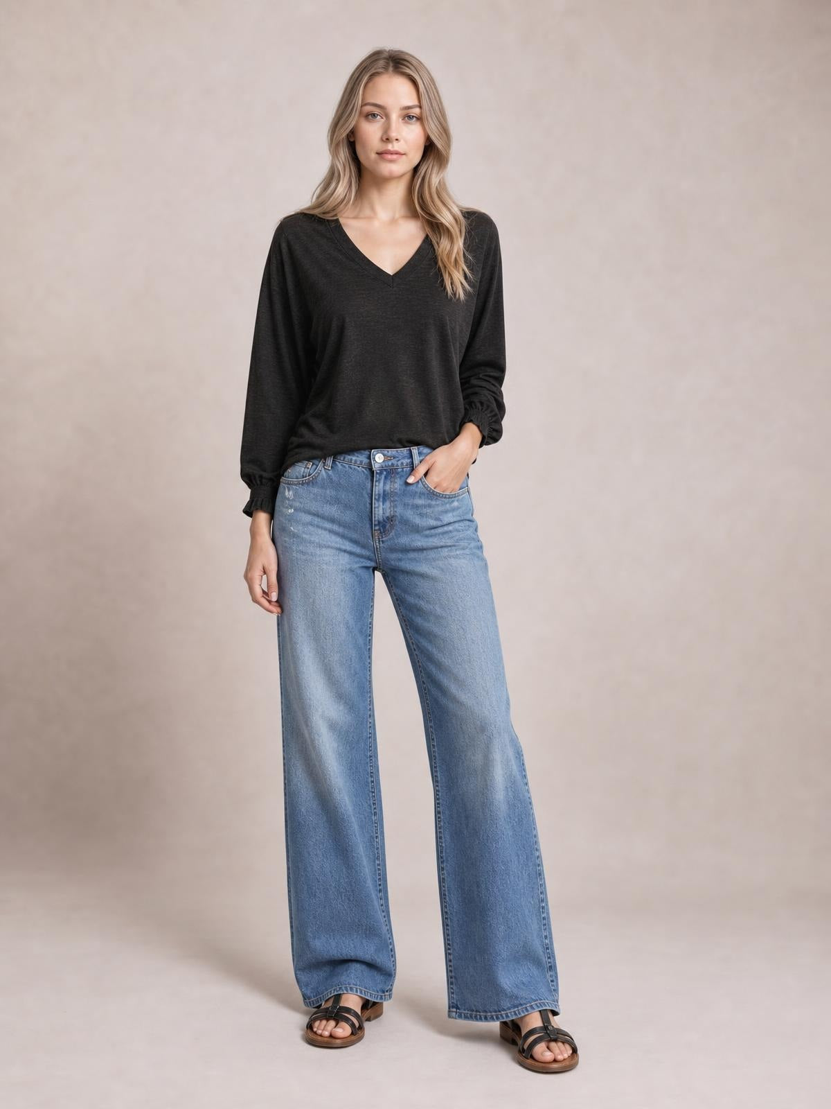Model wears a black notched long sleeve T-shirt with a V-neck, relaxed silhouette, and delicate ruffled cuffs, posing with one hand in her jeans pocket.