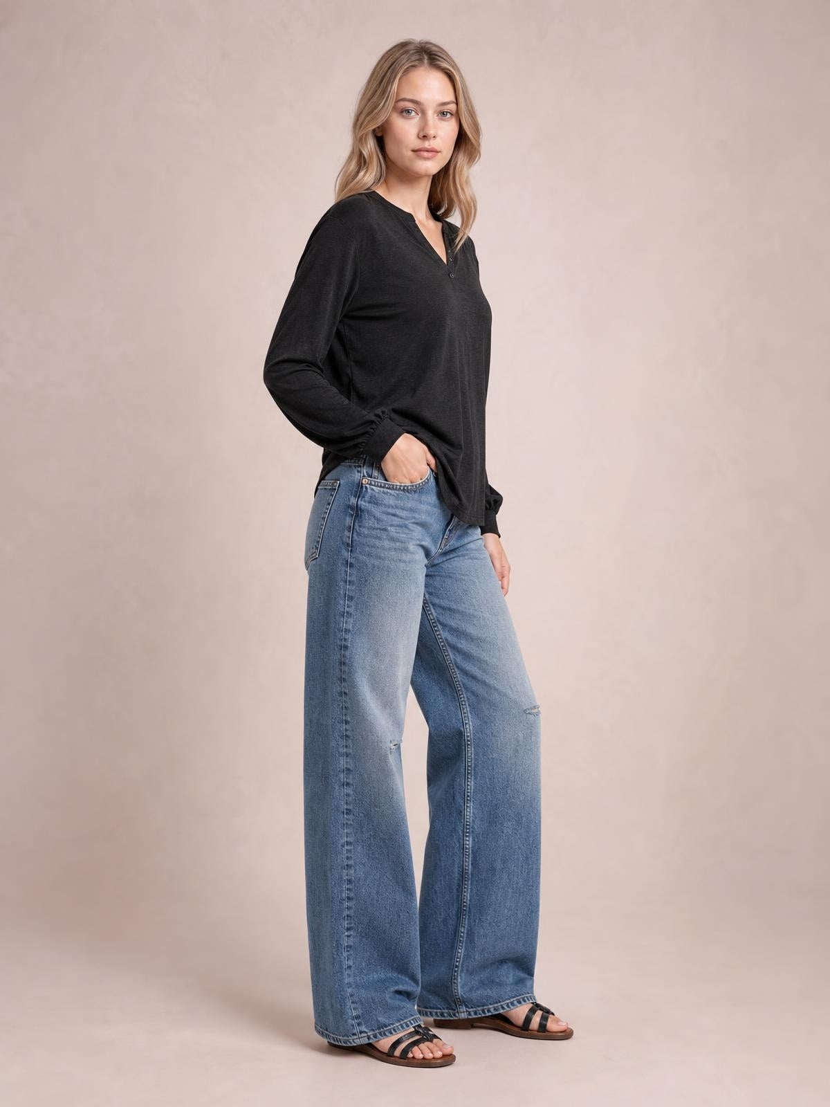 A model poses with one hand in her jeans pocket, wearing a black notched long sleeve T-shirt with a relaxed silhouette and subtle ruffled cuffs.