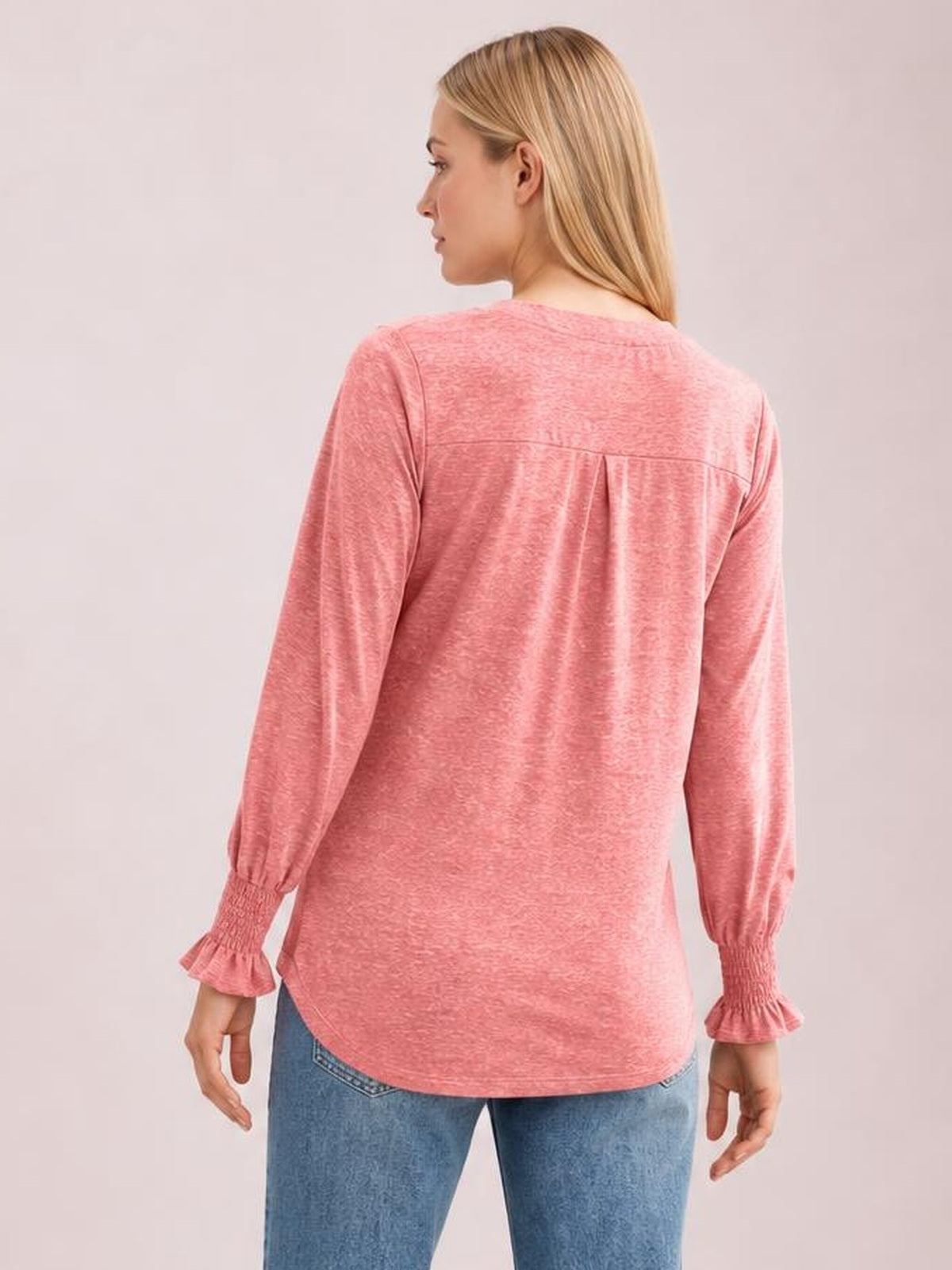 A woman models a pink long sleeve T-shirt from the back, showing a relaxed tunic silhouette, gathered back yoke, and delicate ruffled cuffs.