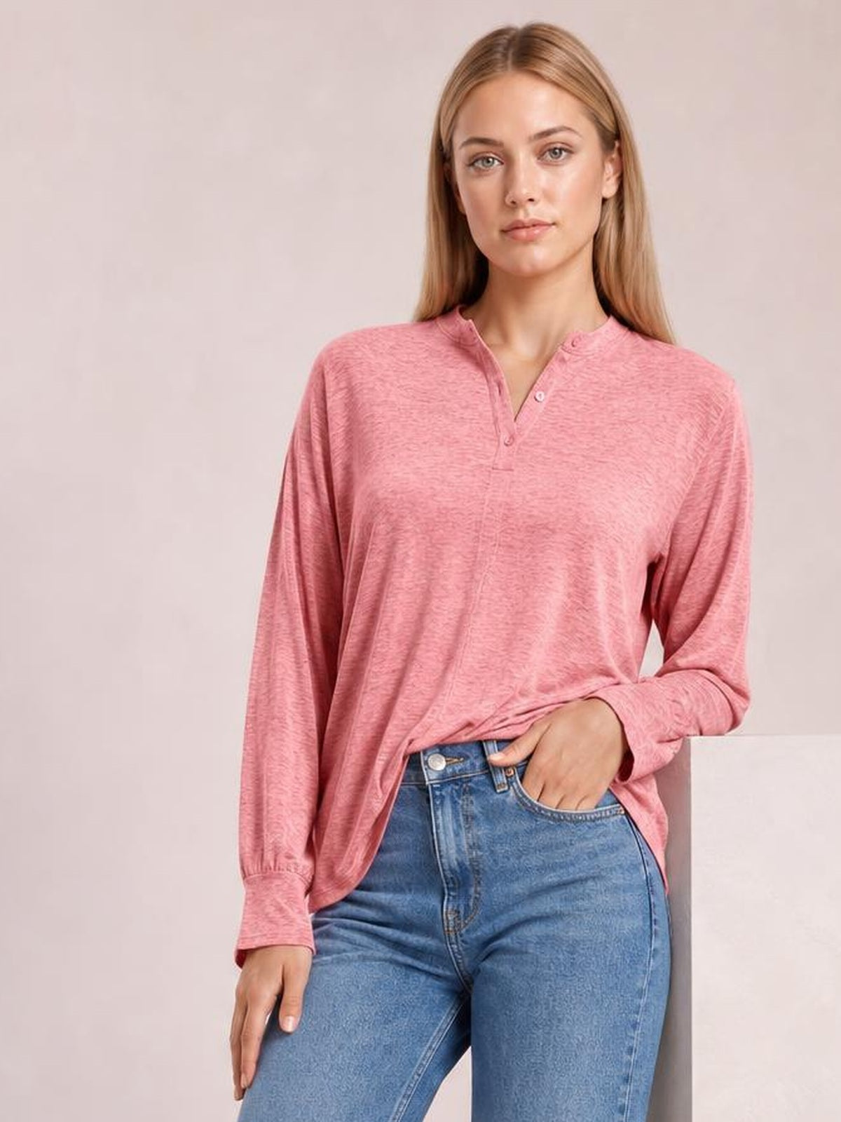 A model wears a pink notched long sleeve T-shirt with a button-front neckline, relaxed silhouette, and slightly gathered cuffs while posing with one hand in her jeans pocket.