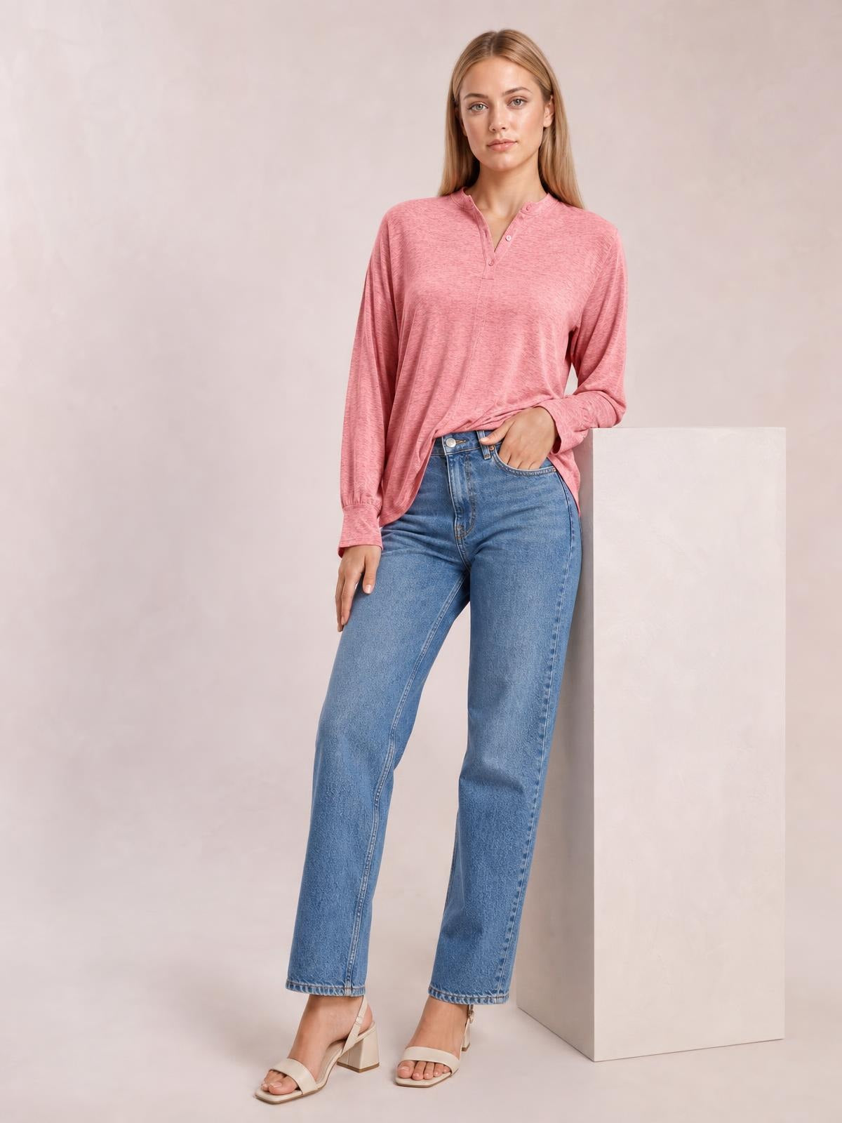 A model wears a pink notched long sleeve T-shirt with a button placket, relaxed silhouette, and straight hem, posing with one hand in her jeans pocket.