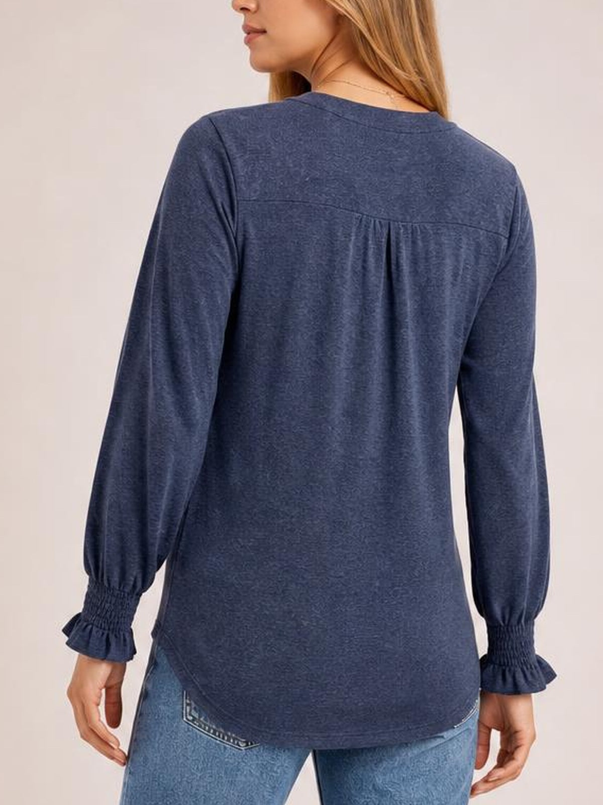A model stands with her back to the camera wearing a navy notched long sleeve T-shirt with a relaxed silhouette, gathered back yoke, ruffled cuffs, and curved hem.