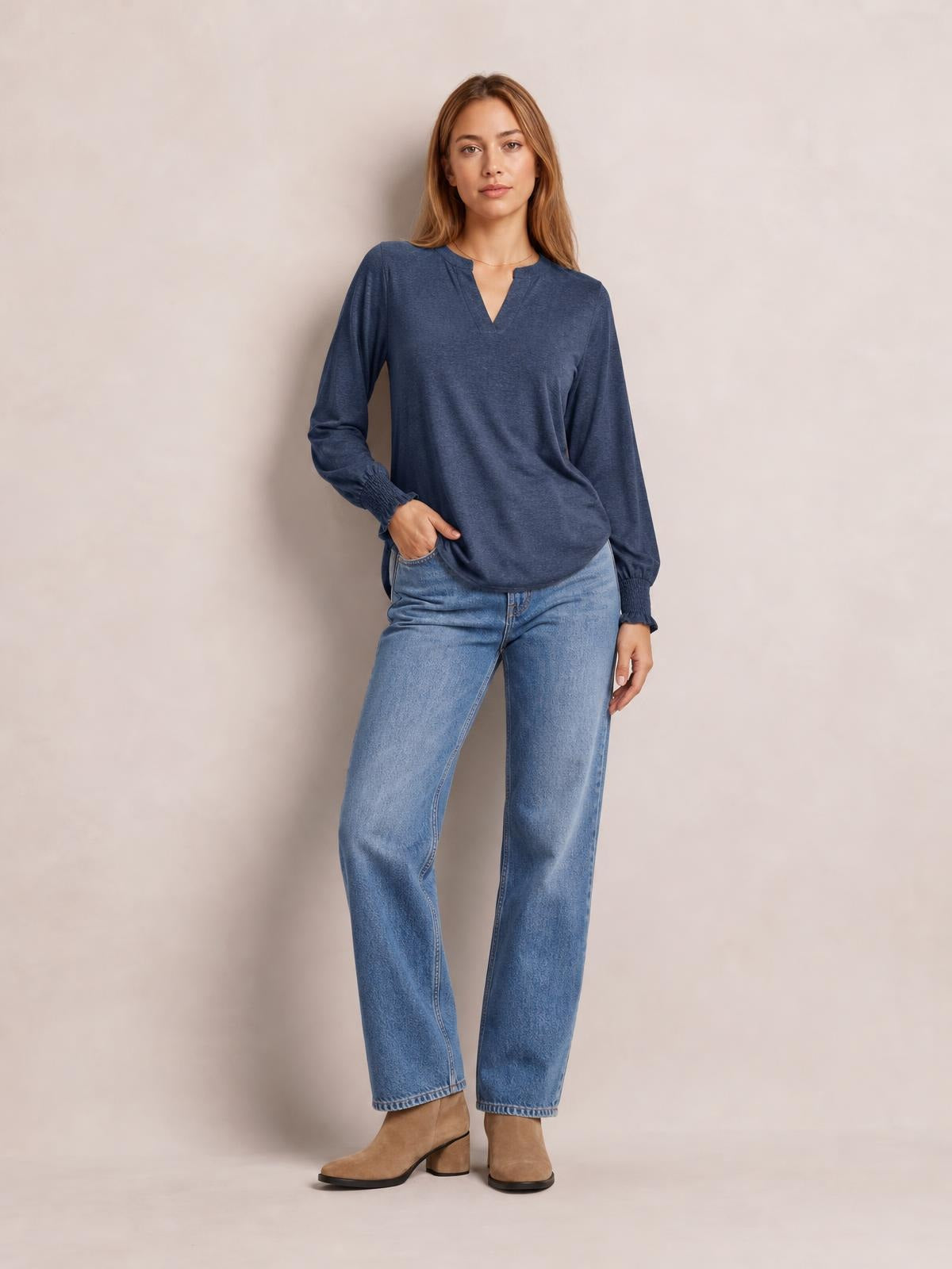 A woman models a navy notched long sleeve T-shirt with a relaxed silhouette, subtle ruffled cuffs, and a slightly curved hem while standing with one hand in her pocket.