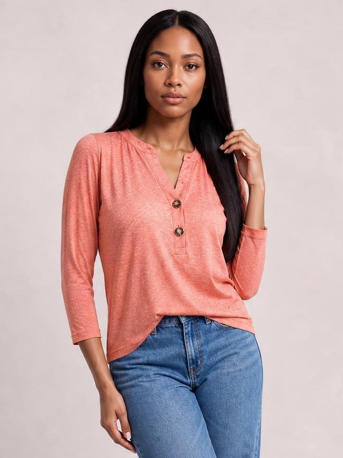 Model poses in a coral pink notched long sleeve T-shirt with a V-neckline, button placket, relaxed silhouette, and softly curved hem.