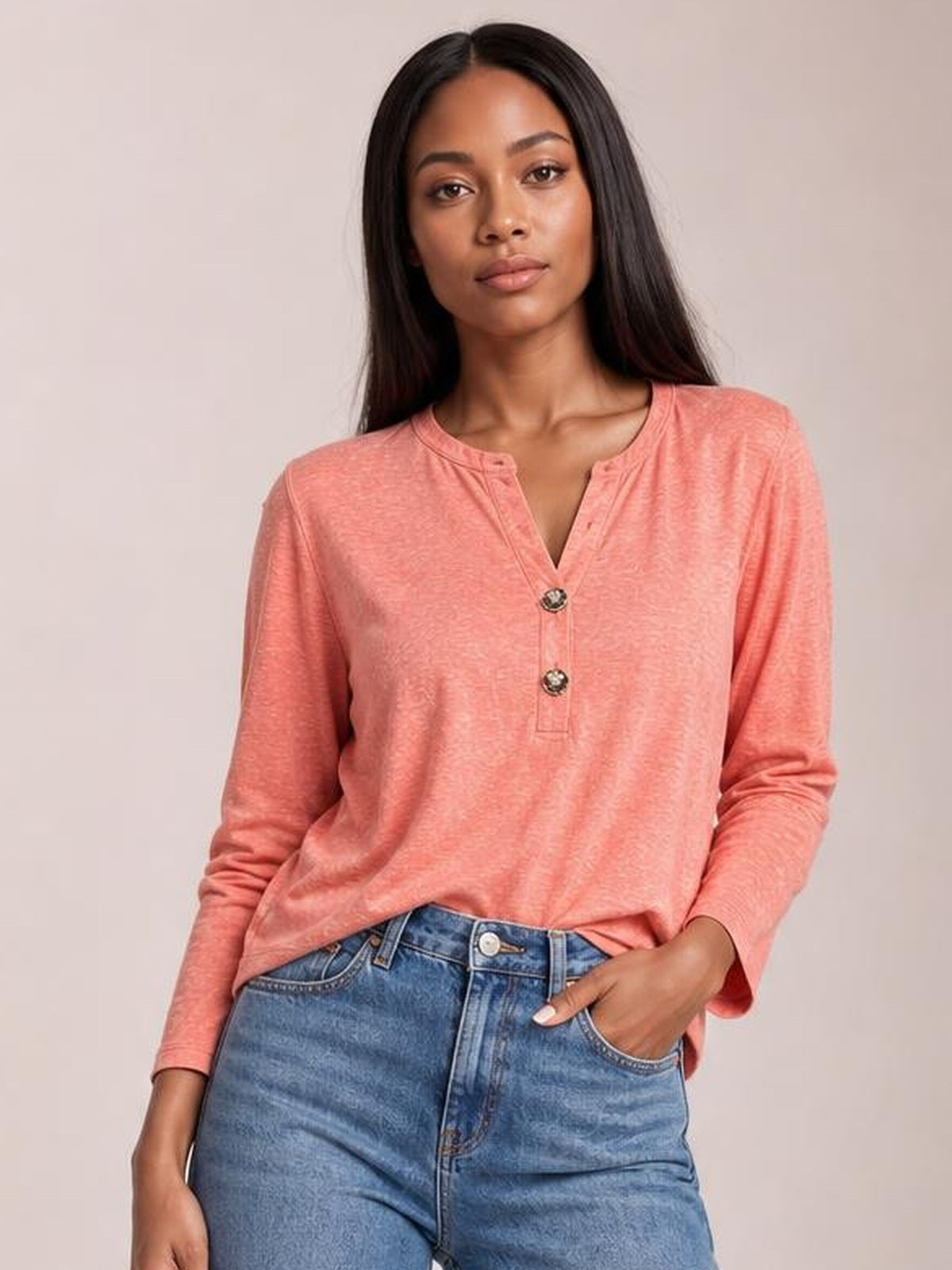 A model wears a coral pink notched long sleeve T-shirt with a relaxed silhouette, round neckline, and two-button placket, posing with one hand in her jeans pocket.