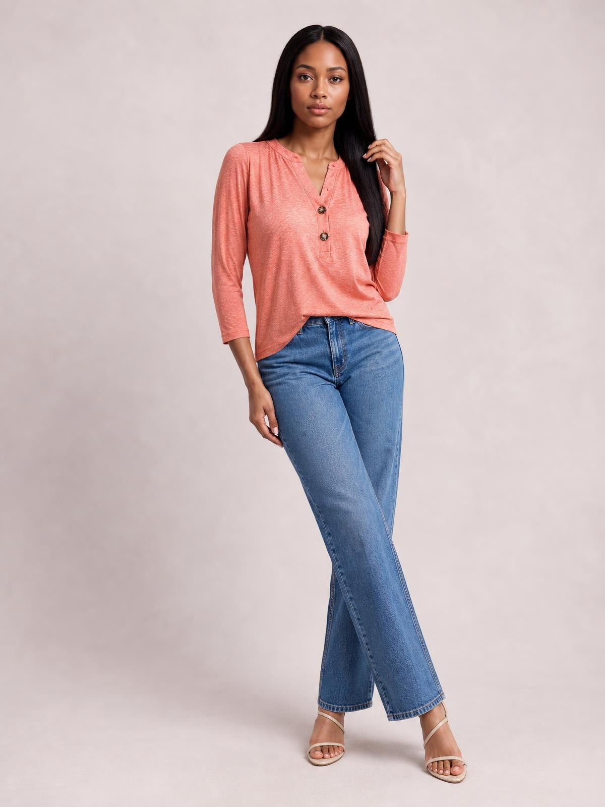 Model wears a coral notched long sleeve T-shirt with a relaxed silhouette, V-neckline, and two-button placket, posing with one hand in her hair.