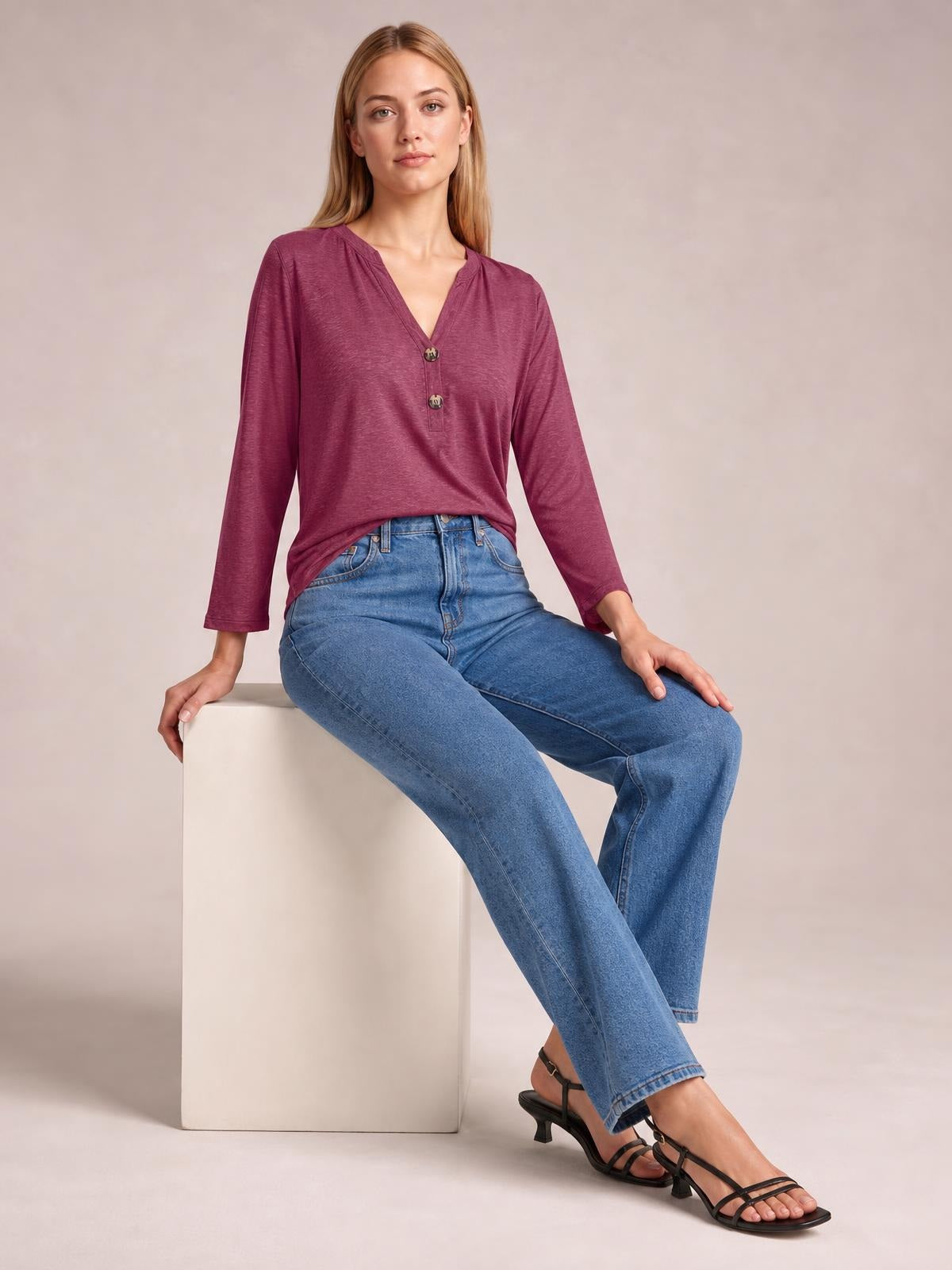 A seated model wears a burgundy notched long sleeve T-shirt with a V-neckline, buttoned front, and relaxed silhouette styled with jeans.